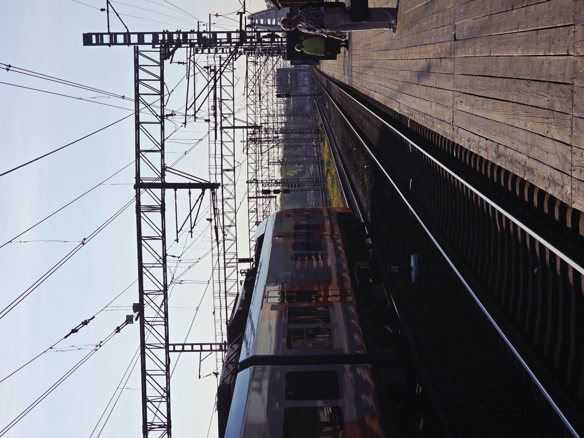 The image shows a railway station in Khimki, Russia. A train is present on the tracks, adjacent to a wooden platform where two people are standing. Overhead power lines and supporting structures are visible above the train and tracks. The photograph is taken at an angle.