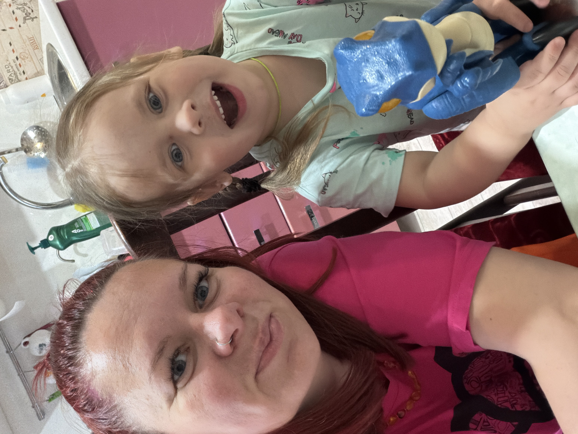 The image is a selfie taken indoors, seemingly in a bathroom or near a wash area, judging by the presence of a sink and toiletries in the background. The foreground is dominated by two individuals: a woman and a young girl. The woman, positioned closer to the bottom of the frame, has reddish-brown hair, fair skin, and blue eyes. She wears a pink top and has a nose ring. The young girl, located at the top of the frame, has blonde hair, blue eyes, and is wearing a light blue shirt with cartoon cat prints. She is holding a blue and cream-colored toy. They are looking directly into the camera, suggesting it's a selfie taken by one of them. The background contains a white sink, a green bottle of liquid soap, pink storage boxes, and an indeterminate chair or furniture. The location may be Chaikovsky, Russia.