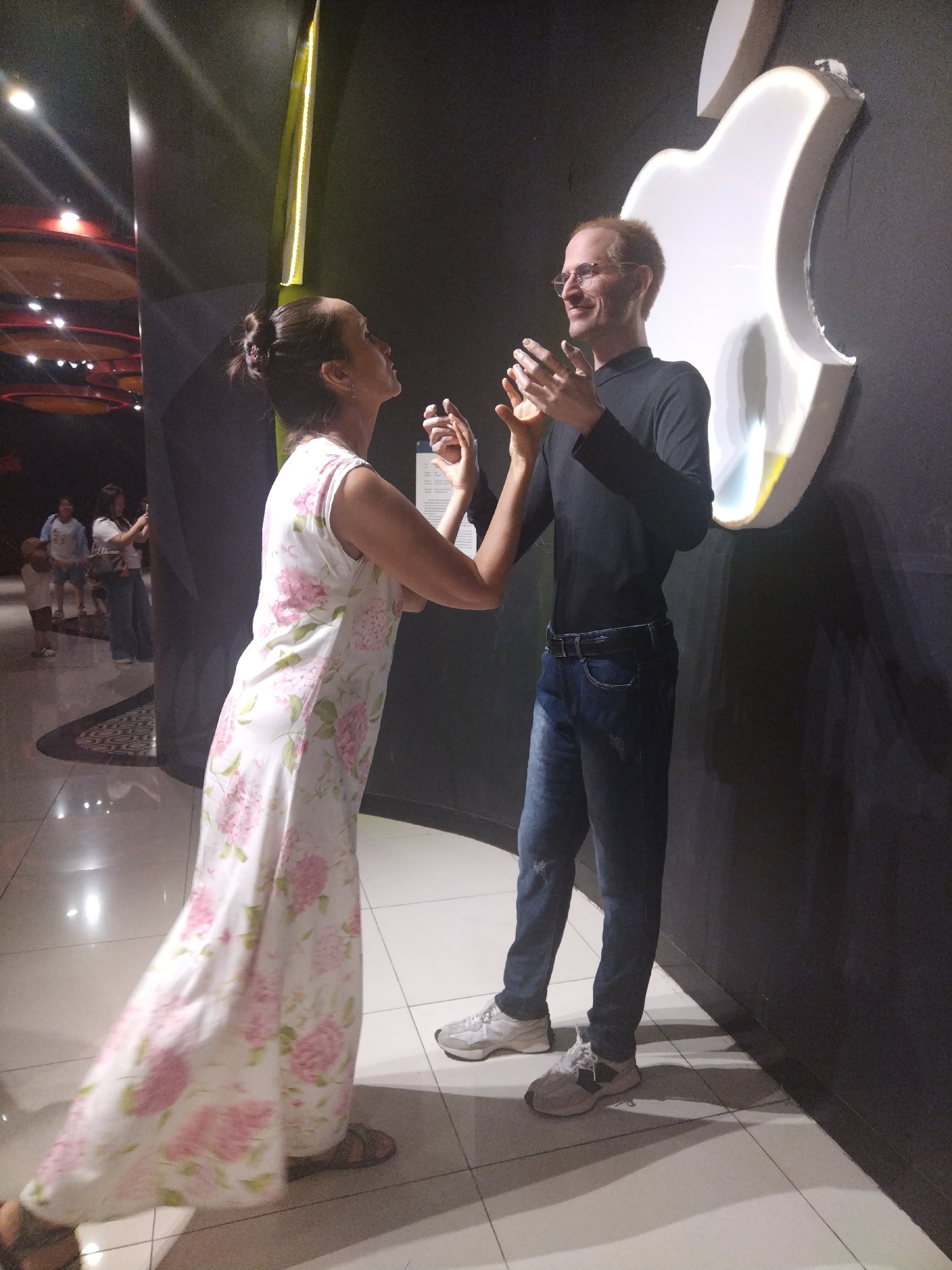 A woman in a floral dress stands with her hands raised near a wax figure of Steve Jobs with a silver Apple logo behind him, inside what looks like a wax museum. Other visitors and museum features are visible in the background. The museum's location is Unknown, Unknown.