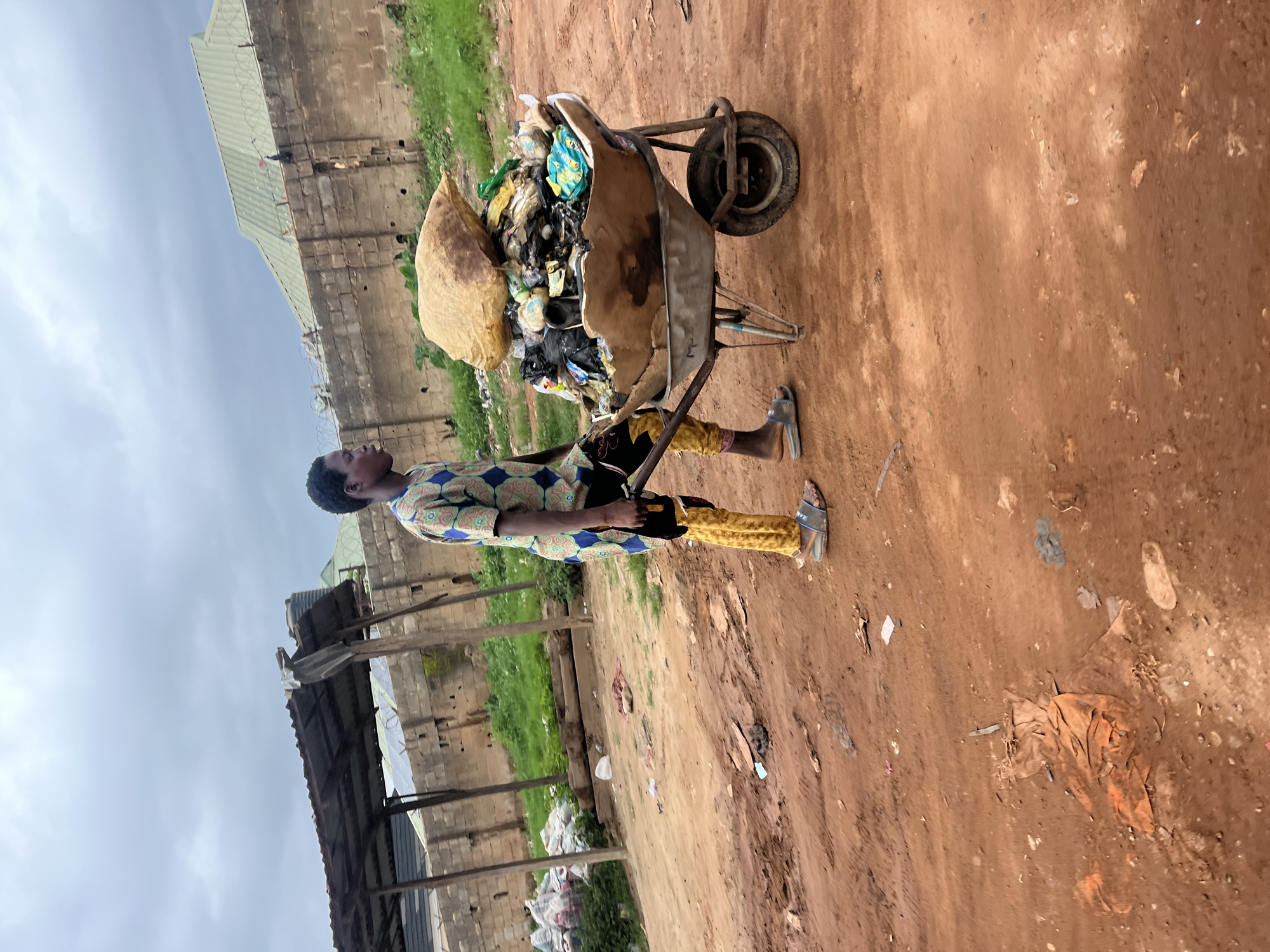 The image displays an individual standing next to a heavily laden wheelbarrow filled with trash, set against a backdrop of a dirt ground and basic buildings under an overcast sky.
The central figure, an individual (likely male), is positioned on the left side of the frame, viewed in profile. They are dressed in a short-sleeved, patterned shirt featuring blue, green, and white geometric designs, paired with yellow patterned trousers. Light-colored sandals are on their feet, with the text "ZAVZ" faintly visible on one.
To the right of the person, a sturdy wheelbarrow is piled high with a large volume of assorted refuse, including numerous plastic bags, wrappers, and general waste items. A significant light-brown sack is also stacked on top of the trash in the wheelbarrow, emphasizing the substantial load.
The ground is a reddish-brown dirt, appearing somewhat uneven and scattered with small pieces of debris. In the background, simple structures made of concrete blocks are visible, some topped with corrugated metal roofs. Patches of green vegetation grow near these buildings. The sky above is uniformly grey and overcast, suggesting a cloudy day. The scene depicts an activity involving the collection or transport of waste, likely within a community context in Abuja, Nigeria.