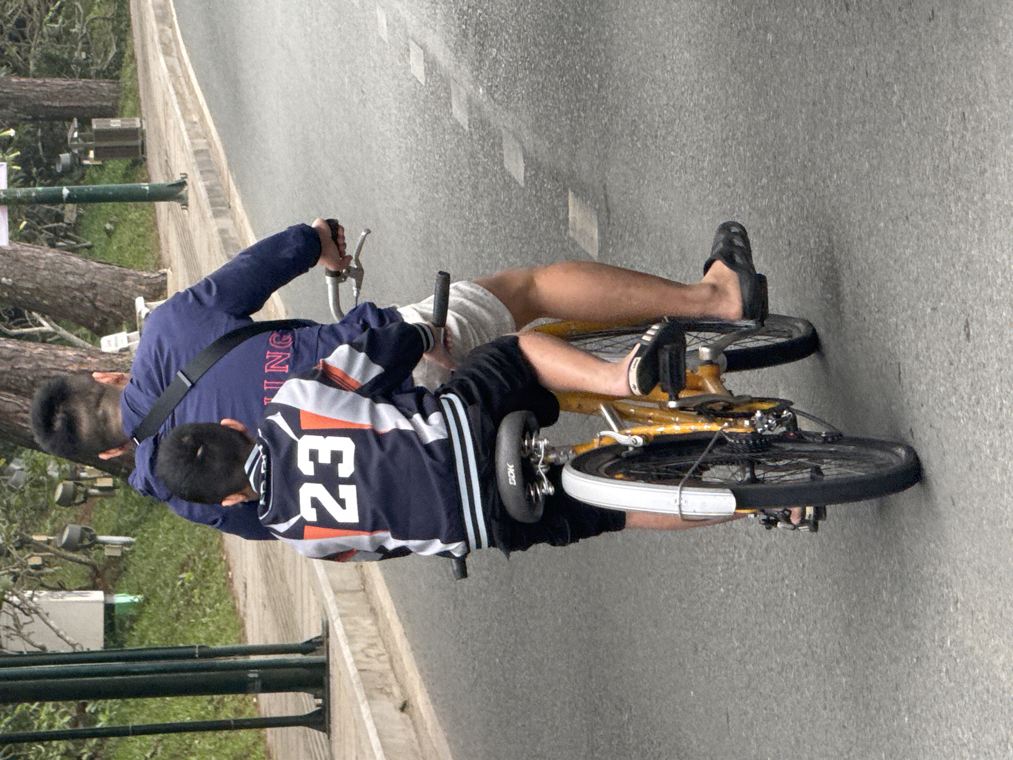 A father and son are riding a bicycle on a paved road in Da Lat, Vietnam. The father is in the front, wearing a dark blue jacket and white shorts. The son is in the back, wearing a blue and white jacket with the number "23" on it, and black shorts. The bicycle is yellow. The road is grey and appears to be a street in a residential area, with trees and buildings visible in the background. The father's hands are on the handlebars, and the son's arms are around his father's waist. The overall atmosphere is one of a typical family outing.