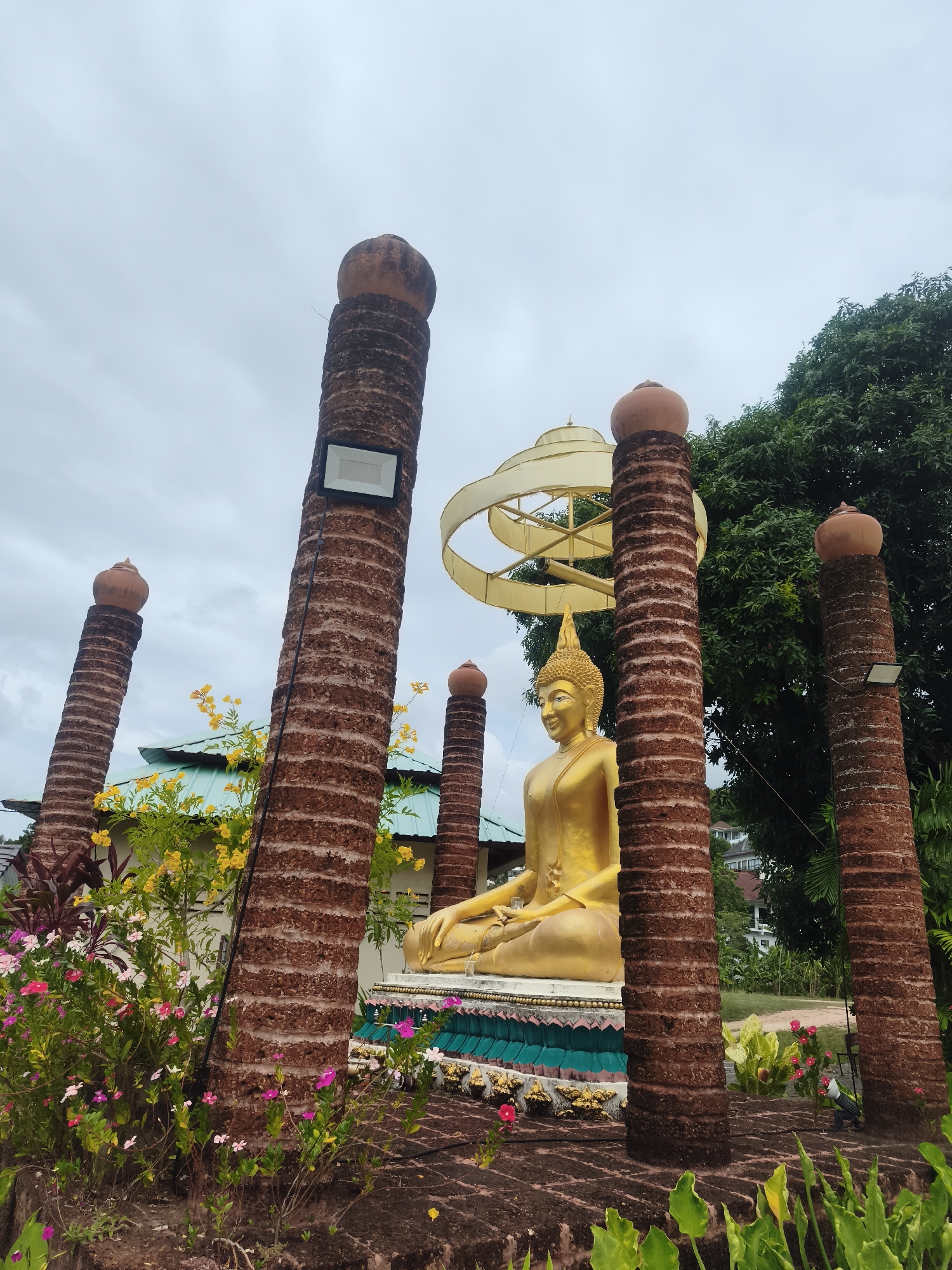 This image depicts a serene outdoor scene, likely a temple or spiritual site in Baan Tai, Thailand. The central focus is a large, golden Buddha statue seated in a meditative posture. The Buddha's face exhibits a gentle, serene expression, and it wears a pointed golden crown or finial. Behind the Buddha's head, an intricate, multi-layered golden halo-like structure is visible.
The Buddha rests upon a decorative, multi-tiered pedestal adorned with teal, white, and golden patterns. Surrounding the statue are several tall, reddish-brown pillars with a rough, textured surface, possibly made of laterite or a similar stone. These cylindrical pillars are topped with spherical, urn-like caps and frame the Buddha, creating a sense of sacred space. A modern floodlight is attached to one of the prominent pillars on the left.
The foreground and mid-ground are populated with lush tropical foliage and vibrant flowers, including pink, yellow, and red blooms, suggesting a well-maintained garden. In the background, large green trees provide a natural setting. Parts of a building with a light blue or teal roof are visible behind the plants on the left, and further in the distance, other structures or residences can be seen on a hillside. The sky is overcast and cloudy, indicating daytime with diffused natural light. There is no clearly legible text visible on any of the objects in the image.