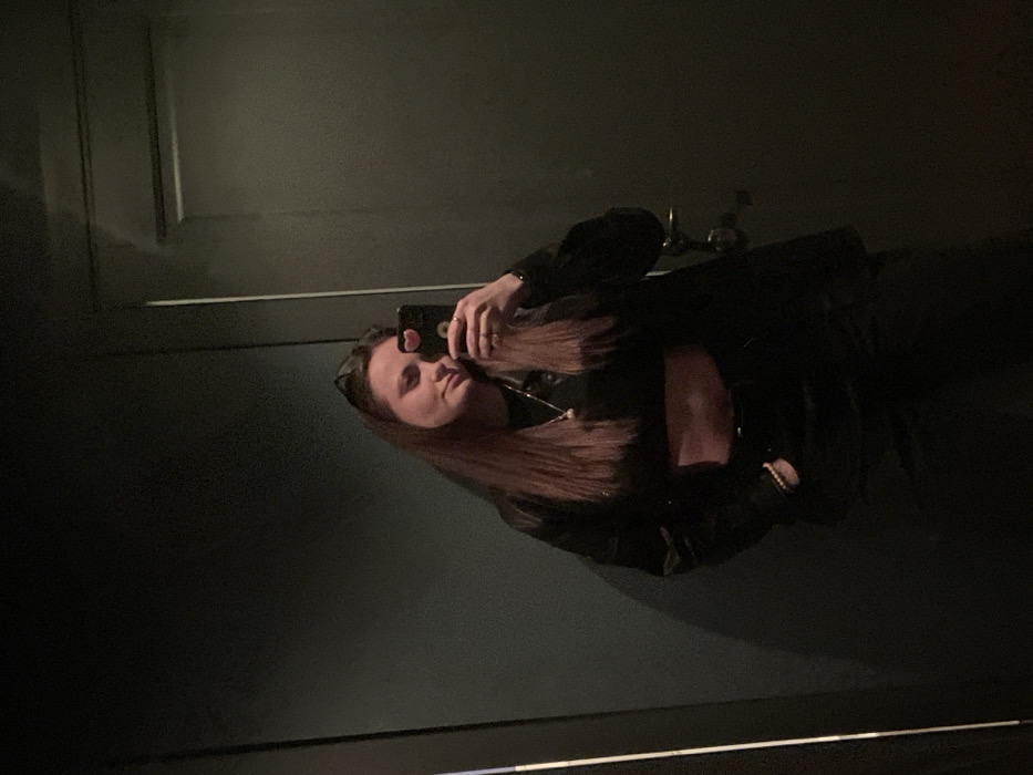 A woman takes a selfie in a dark, reflective space, possibly a mirror. She wears a black jacket and top, and her long, light brown hair is visible. Her phone is held up in her hand. The background is a dark, plain surface with what appears to be a door frame in the upper left corner and a sink with a faucet in the top center, suggesting she may be in a bathroom. The lighting is dim.