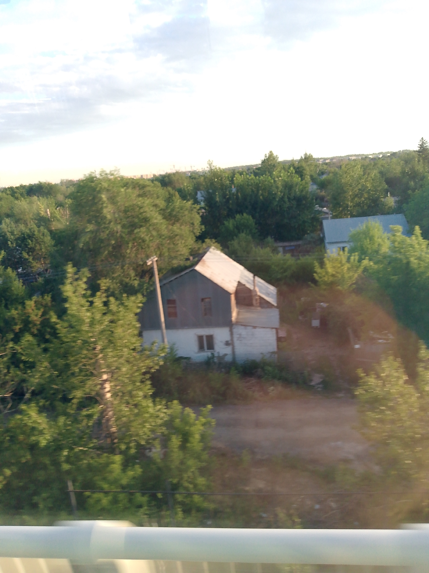 The image depicts a rural landscape with a two-story house nestled amongst lush green trees. The house has a white lower section and a darker, possibly gray or metal, upper section with a pitched roof. Two windows are visible on the upper level, and one on the lower level. A wooden utility pole stands near the house. In the background, a larger, more modern-looking building with a light-colored roof is partially visible, also surrounded by trees. The scene is bathed in the warm, golden light of what appears to be either early morning or late afternoon, suggesting a clear or partly cloudy sky. The overall impression is one of a tranquil, natural setting, likely in a suburban or rural area. The image appears to have been taken from a moving vehicle, as indicated by the blurred white horizontal element at the bottom of the frame. No people are visible in the image, and there is no discernible text.