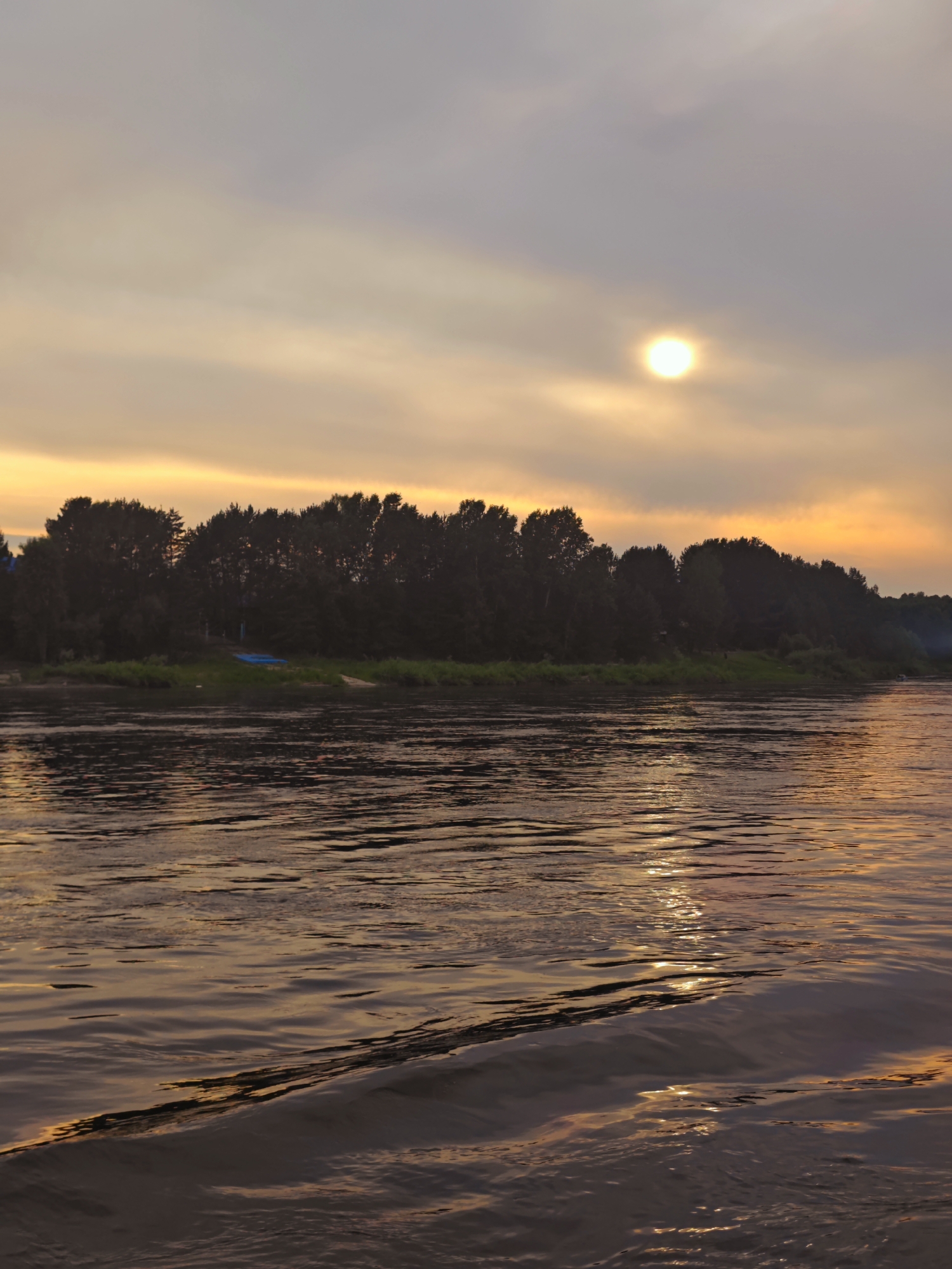 A sunset scene over a wide river, viewed from the water's surface. The sky is filled with diffused sunlight and soft clouds, tinged with yellow and orange hues, suggesting dusk. A bright, circular sun is visible in the upper right portion of the sky, partially obscured by clouds. The opposite bank of the river is lined with a dense, dark silhouette of trees. On the bank, a small, blue object, possibly a boat or cover, rests on the grassy shore. The river's surface is rippled, reflecting the colors of the sky and creating streaks of light. In the foreground, the water shows distinct wakes and wave patterns. The overall atmosphere is serene and natural, indicative of a rural or wilderness setting near YUKSEEVO, RUSSIA.
