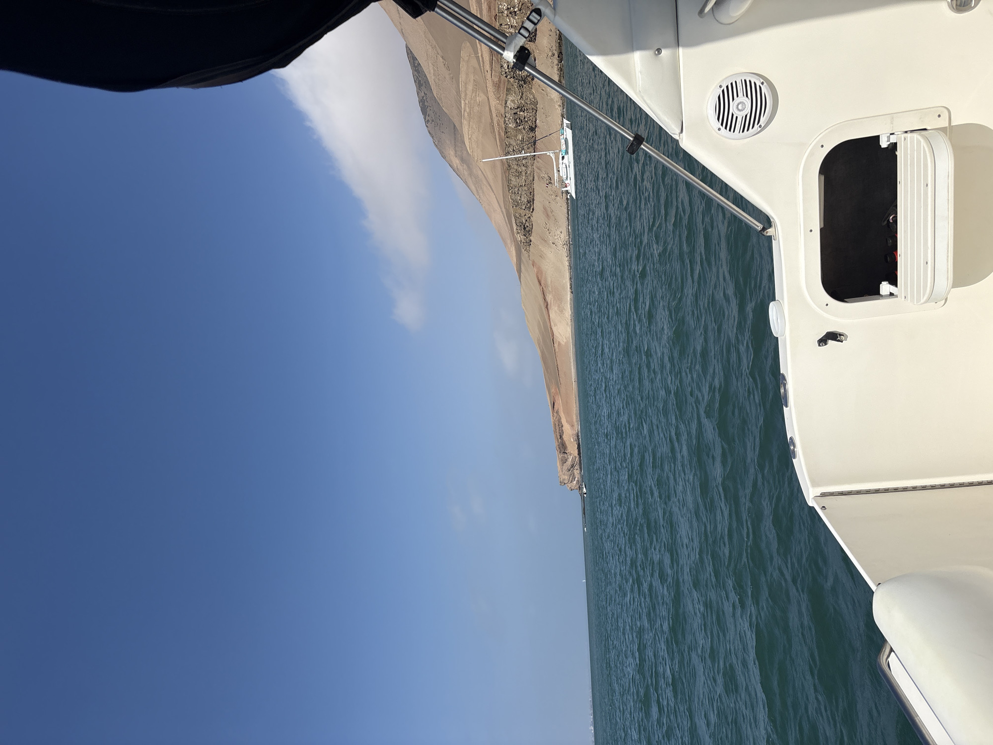 View from a boat at sea, presumably off the coast of Callao, Peru, as evidenced by the arid coastal terrain visible. The image captures the ocean, a clear blue sky with light clouds, and a stretch of shoreline. A catamaran-type sailboat is anchored near the shore. Part of the boat from which the picture is taken is in the foreground, with metallic support bars and a compartment door ajar. The ocean is dark blue-green.