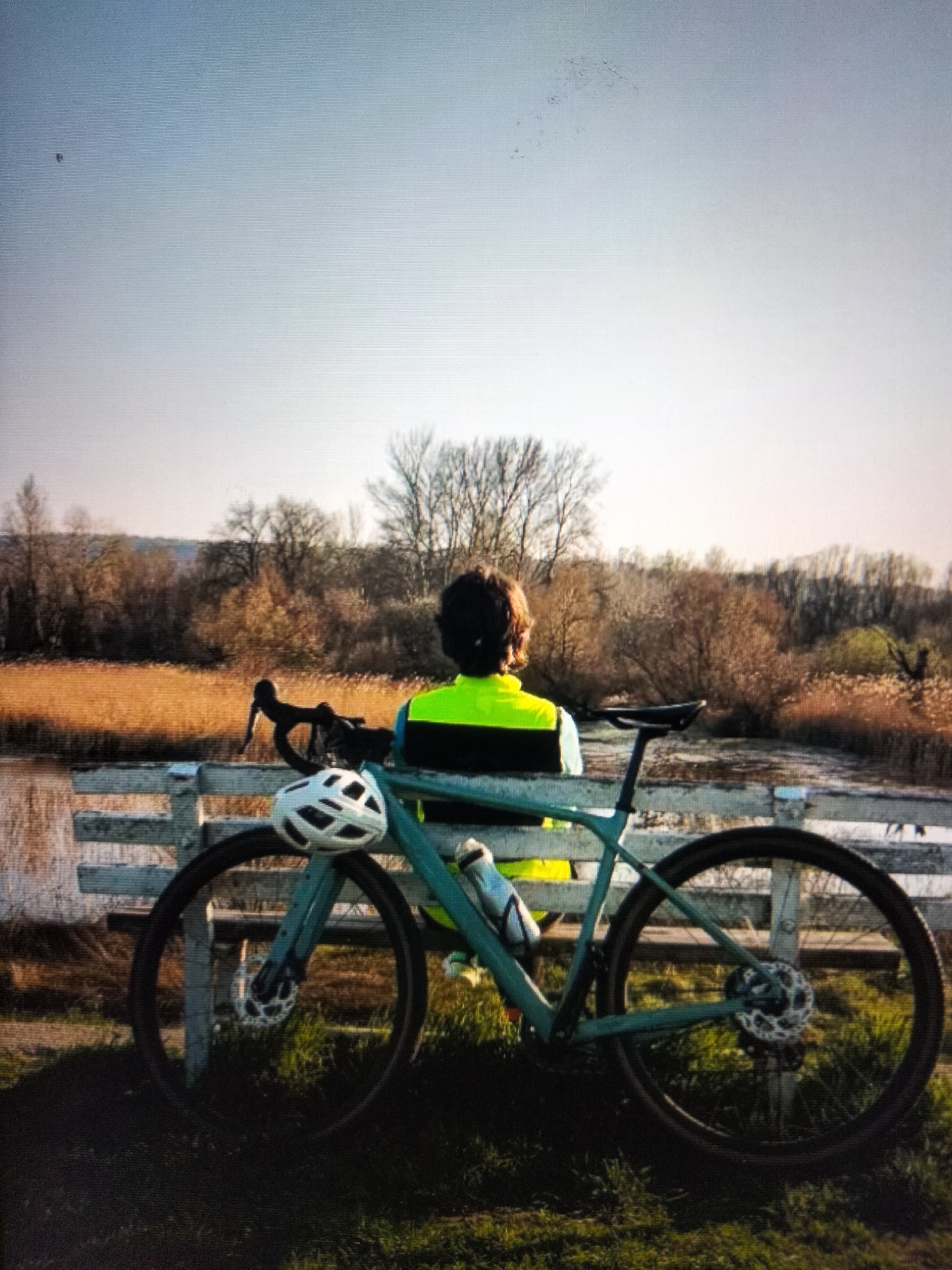 A person wearing a bright yellow and black cycling jersey and a white helmet is seated on a light blue bicycle. The bicycle has a water bottle attached to the frame. The person and bicycle are positioned in front of a white wooden fence. Beyond the fence is a body of water and a landscape with dry reeds and trees. The sky is clear and light blue, with a gradient towards white at the horizon. The scene is outdoors, likely in a park or natural area.
