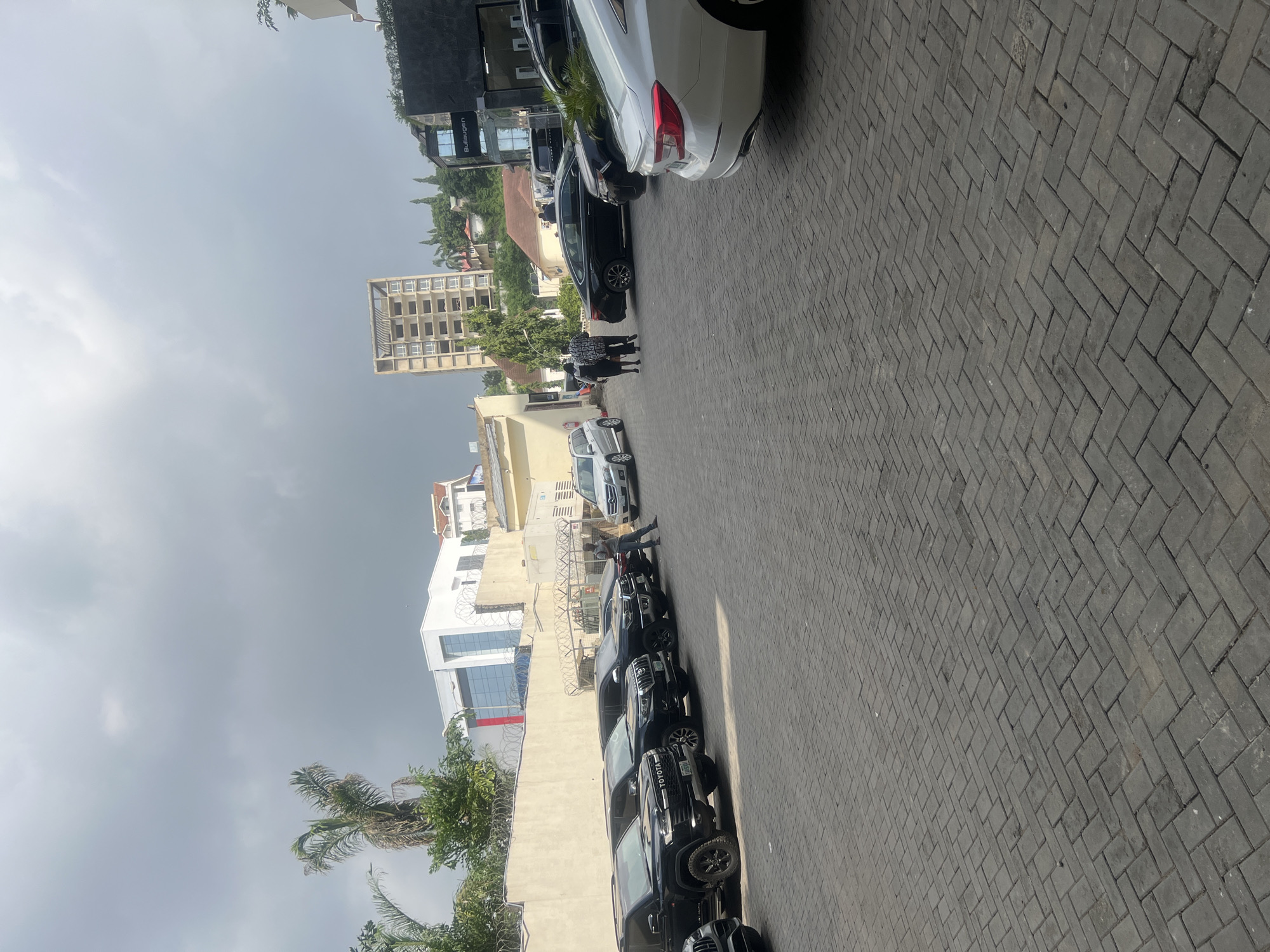 This image depicts an outdoor scene with several cars parked along a paved walkway. The cars, predominantly black SUVs, are lined up in a row. A few people are visible, with one individual interacting with a car and others walking in the distance. In the background, there are buildings of varying architectural styles, including modern structures with large glass windows and older, more traditional designs. Some greenery, including palm trees and other foliage, is interspersed with the buildings. The sky is overcast, suggesting a cloudy or possibly humid day. The paving stones on the walkway are laid in a herringbone pattern. The overall impression is of a business or residential area. A sign on one of the buildings in the distance appears to read "Bujagen." The scene is likely taking place during daylight hours, given the ambient light.