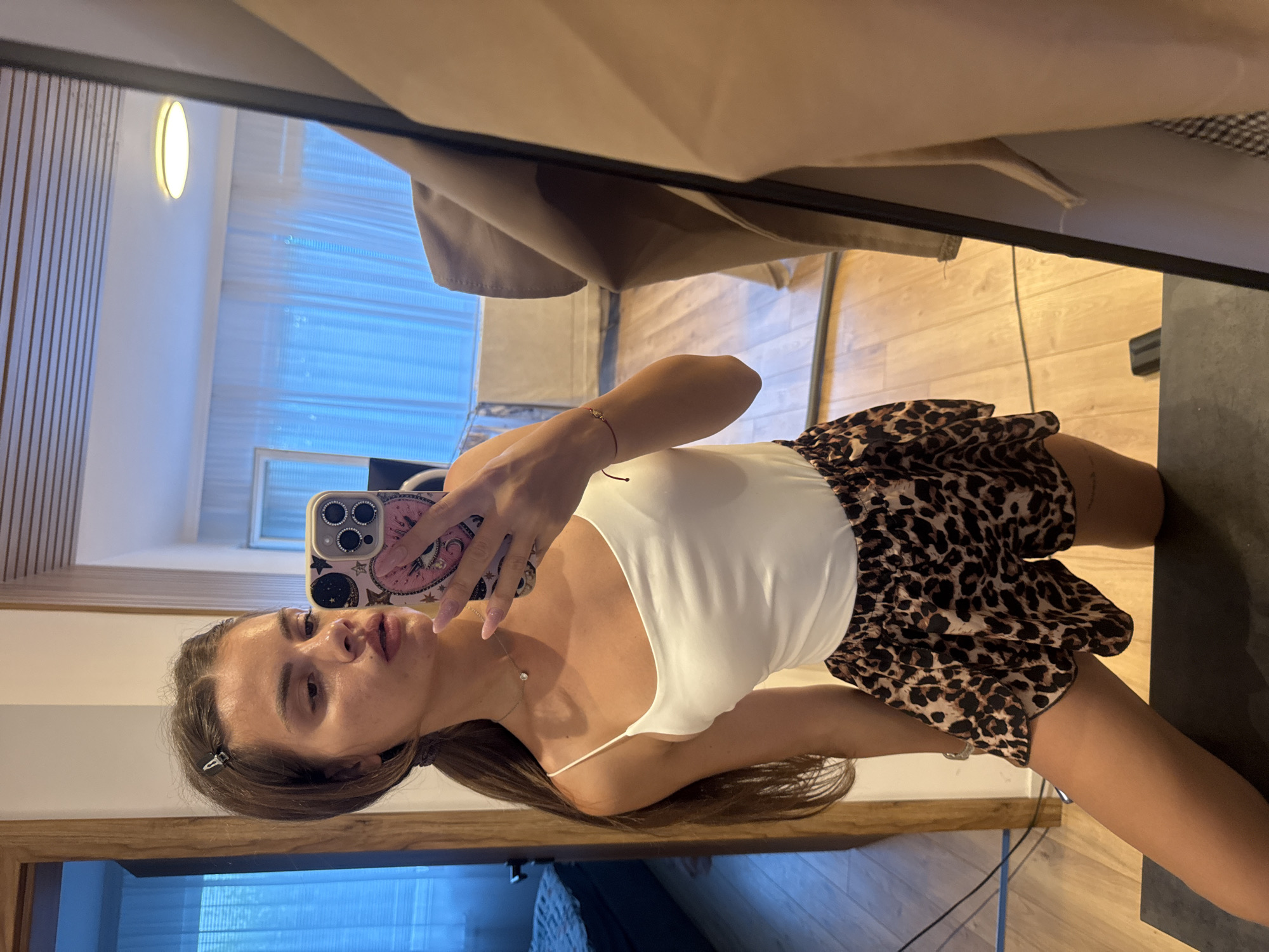 A young woman is taking a mirror selfie in what appears to be a bedroom. She is wearing a white camisole and leopard-print shorts. Her hair is pulled back in a ponytail, and she has a hair clip in the front. She is wearing a necklace and a bracelet. In the background, a mirror reflects the room, showing a bed, a window with blue blinds and sheer curtains, and a ceiling light. The floor is wooden. The image does not contain any information that would indicate the location in the city of Plovdiv, Bulgaria.