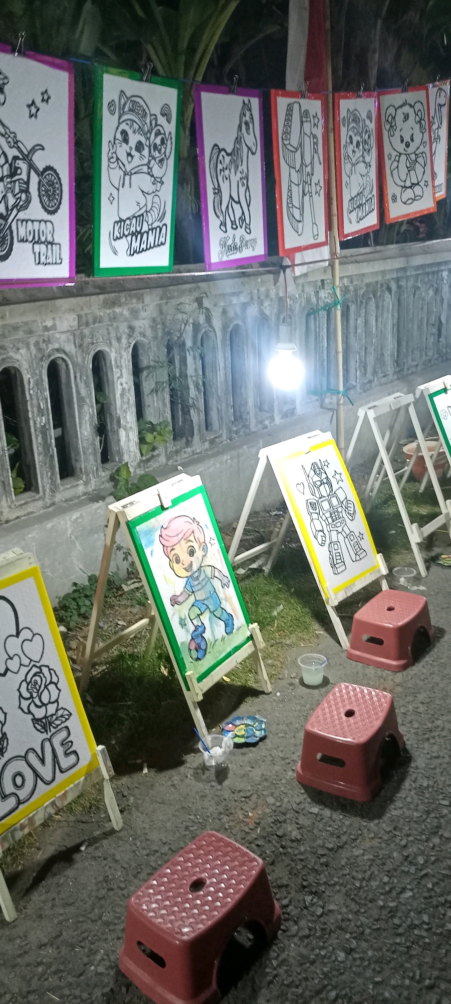 A row of framed coloring pages, featuring cartoonish designs like a motorcycle, cats, a horse, a robot, and a teddy bear, are displayed outdoors at night, likely in Kalimati, Indonesia, possibly at a market or street event. The pages are hung on a wire above a concrete wall with decorative arched openings. Below, three easels stand upright, each displaying a partially colored page. One easel shows a cartoon boy playing soccer, another a drawing of the word "LOVE" with a rose and hearts, and a third a robot. Two small red plastic stools are placed on the asphalt ground near the easels, suggesting a painting activity. A single light bulb hangs overhead, illuminating the scene.