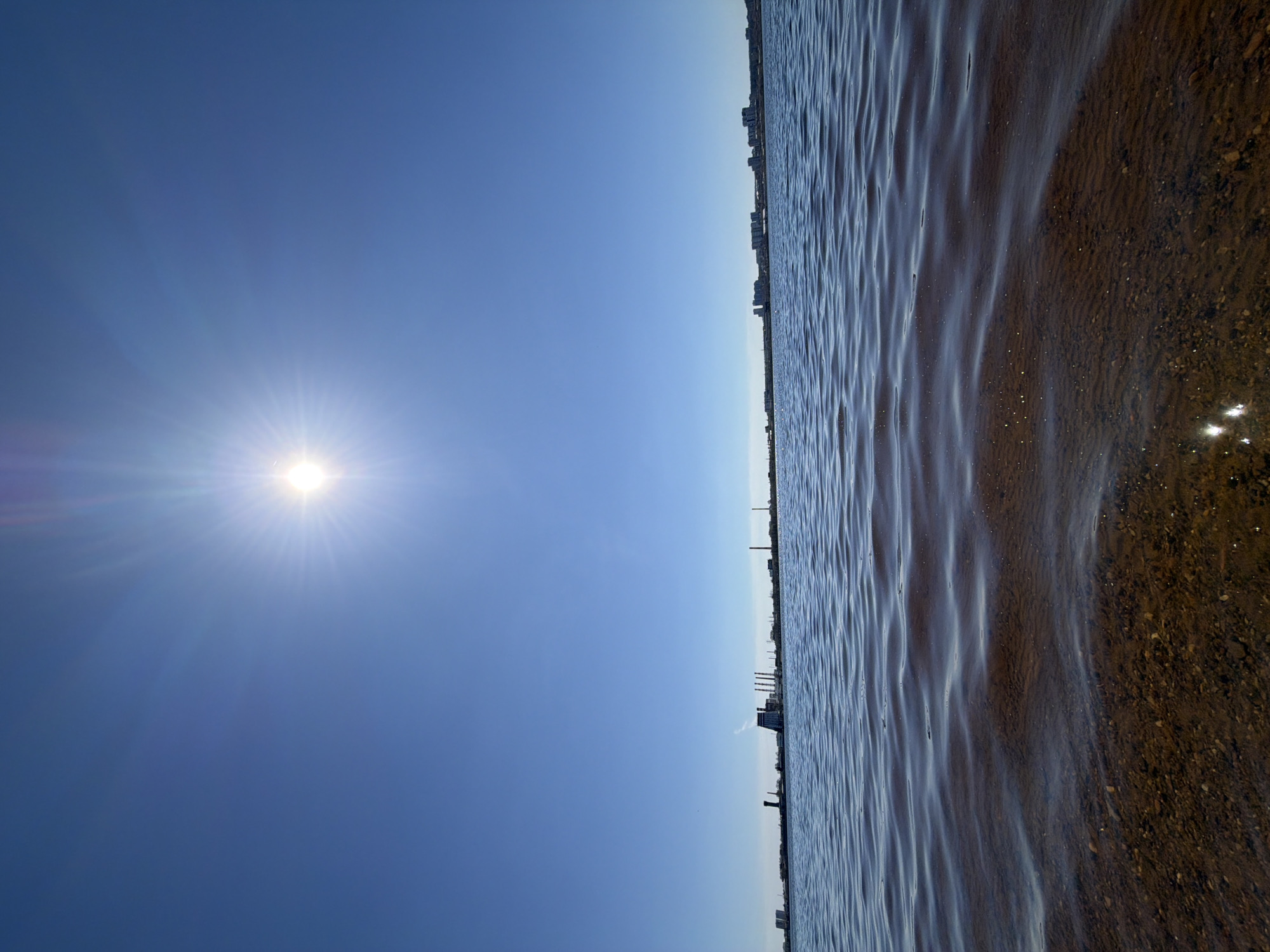 The image depicts a clear blue sky with a bright sun, and a body of water (likely a lake or river) next to it. The sun is positioned in the upper left. The water exhibits small ripples and reflects some sunlight. A coastline with distant buildings, possibly a cityscape, is visible across the water. There are no people visible. The location may be Izhevsk, Russia. The environment is sunny and bright, indicating a clear weather day.