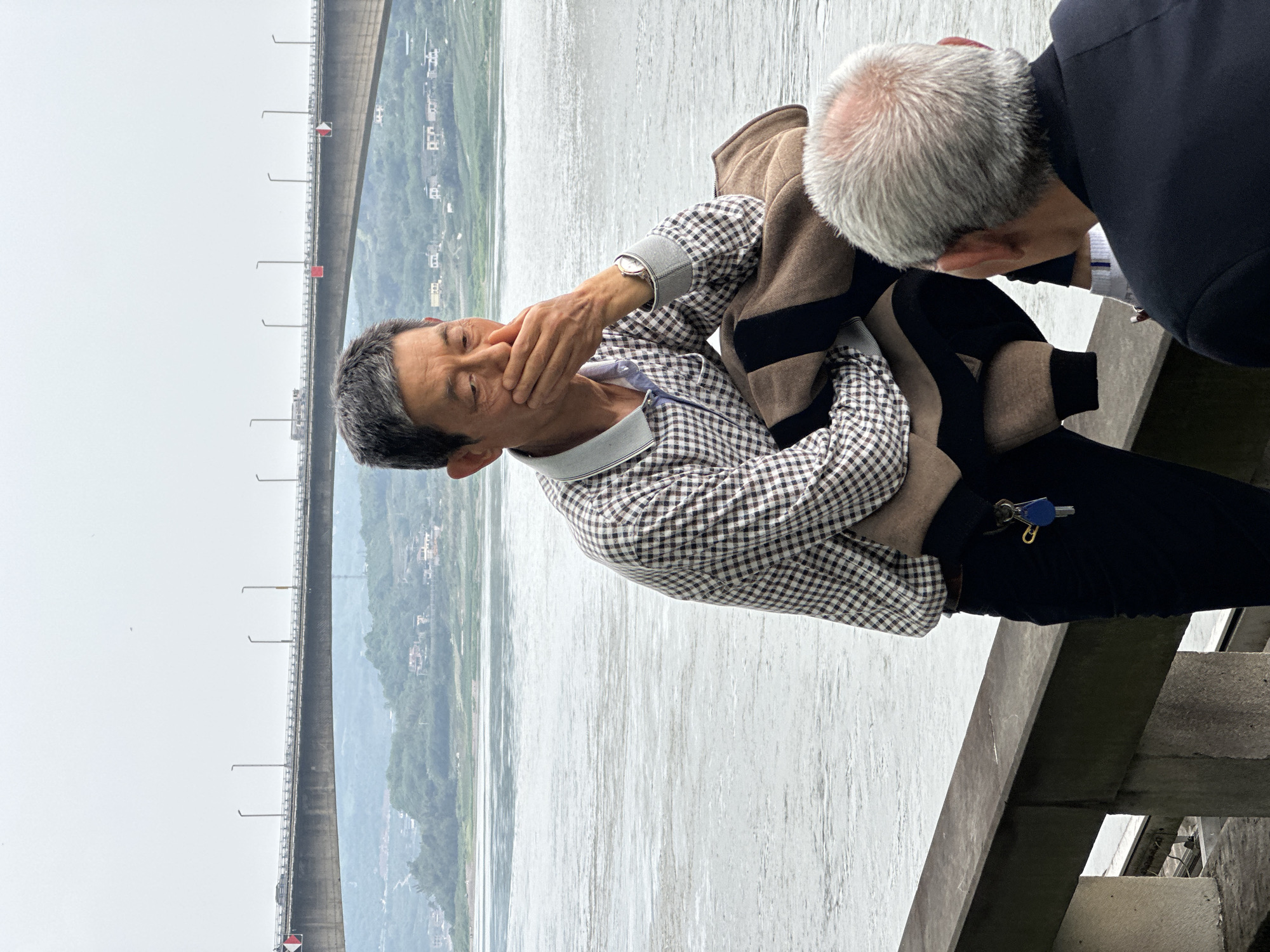 On an overcast day, two older Asian men are seen on a concrete structure, possibly a bridge or dam in Jiangjin, China. One man, with gray hair and wearing a black and white checkered shirt and black pants, is seated with his hand covering his mouth. A jacket with brown and black stripes is draped over his lap. A set of keys hangs from his pants. The second man, wearing a dark suit, is bent over, possibly speaking to the seated man. In the background, a large concrete structure spans a body of water, with hills and buildings visible in the distance. Streetlights line the structure. The scene suggests a somber or private moment between the two men, likely at a tourist attraction.
