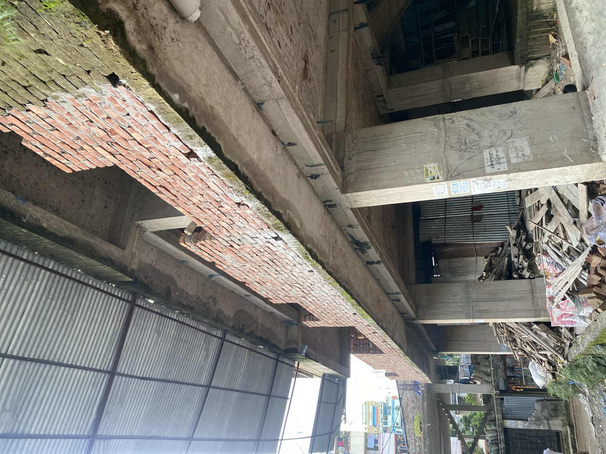 The image depicts a construction site in Dhaka, Bangladesh. The scene is dominated by unfinished concrete structures and exposed brick walls, indicating ongoing building work. A corrugated metal roof extends over part of the area, providing some shelter. Various construction materials, including piles of wood, bricks, and bags of cement, are scattered around the site. The ground appears to be uneven and littered with debris. In the background, a few buildings can be seen, suggesting an urban environment. The overall impression is one of a busy and somewhat disorganized construction zone in a densely populated city.