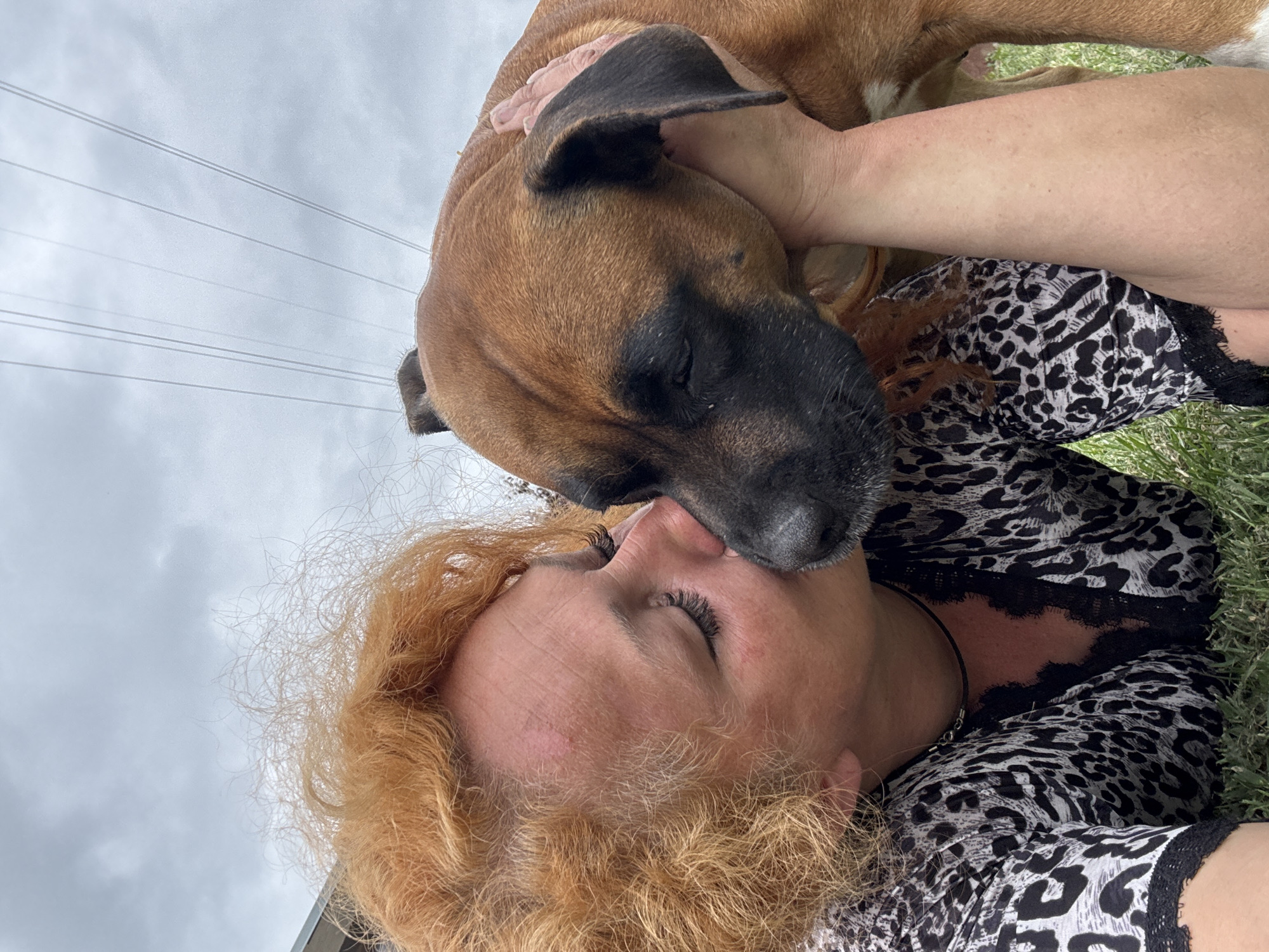A woman with bright red, curly hair and dark eyelashes is captured in a close-up outdoor selfie, leaning in to kiss a tan-colored dog with a black muzzle. The woman is wearing a black and white leopard-print top with lace trim. The dog, held gently by the woman's hand on its head, is positioned to receive the kiss. They are both lying down on green grass, with a cloudy, overcast sky and power lines visible in the background. The scene appears to be a moment of affection between the woman and her pet, likely in a park or backyard setting within the city of Chaikovsky, Russia.