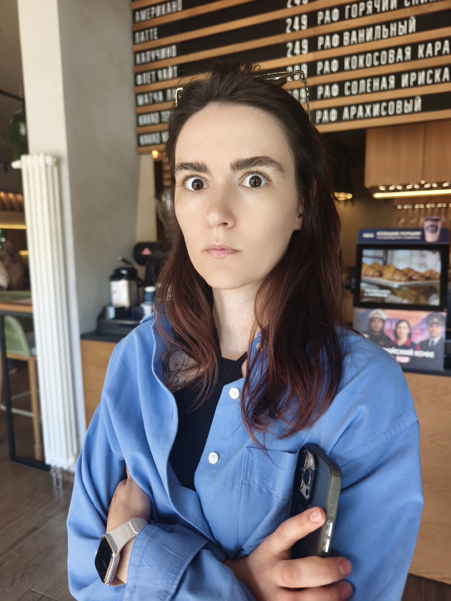 A young woman with long, dark brown hair with reddish undertones is captured in a half-body shot inside a bright cafe in Tyumen, Russia. She has a pale complexion and is looking directly at the camera with a wide-eyed, somewhat surprised or theatrical expression. Her glasses are pushed up onto her head. She is wearing a light blue, long-sleeved button-up shirt over a black top, with a glimpse of a tattoo on her left shoulder. Her arms are crossed, and she is holding a dark-cased smartphone with multiple camera lenses in her right hand, tucked against her left arm. A light-colored smartwatch with a dark screen is visible on her left wrist.
The background reveals a typical cafe setting. Directly behind her is a wooden menu board listing various coffee drinks and their prices in Cyrillic. Notable items include "АМЕРИКАНО" (Americano), "ЛАТТЕ" (Latte), "КАПУЧИНО" (Cappuccino), and several "РАФ" (RAF) flavored coffees such as "ГОРЯЧИЙ" (Hot), "ВАНИЛЬНЫЙ" (Vanilla), "КОКОСОВАЯ КАРА" (Coconut Caramel), "СОЛЕНАЯ ИРИСКА" (Salted Toffee), and "АРАХИСОВЫЙ" (Peanut), many priced at 249. To the right, a display case shows pastries, specifically croissants, and a smaller sign below it features images of people and partial Cyrillic text, including what appears to be "ЧЙСКИЙ КОФЕ" (likely "Czech Coffee"). Part of a coffee machine or equipment is visible in the mid-background. On the far left, a white radiator stands vertically. The lighting suggests it is daytime, contributing to the cafe's inviting atmosphere.