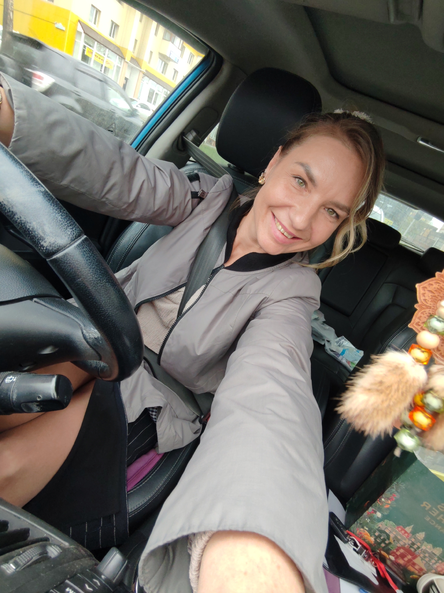 A woman is sitting in the driver's seat of a car, smiling at the camera. She is wearing a light gray jacket over a beige top. Her left arm is extended, resting on the steering wheel. The interior of the car is visible, including black leather seats and the dashboard. Outside the car window, buildings and other vehicles can be seen. The background suggests an urban environment, possibly in Petropavlovsk-Kamchatsky, Russia, based on the provided context.