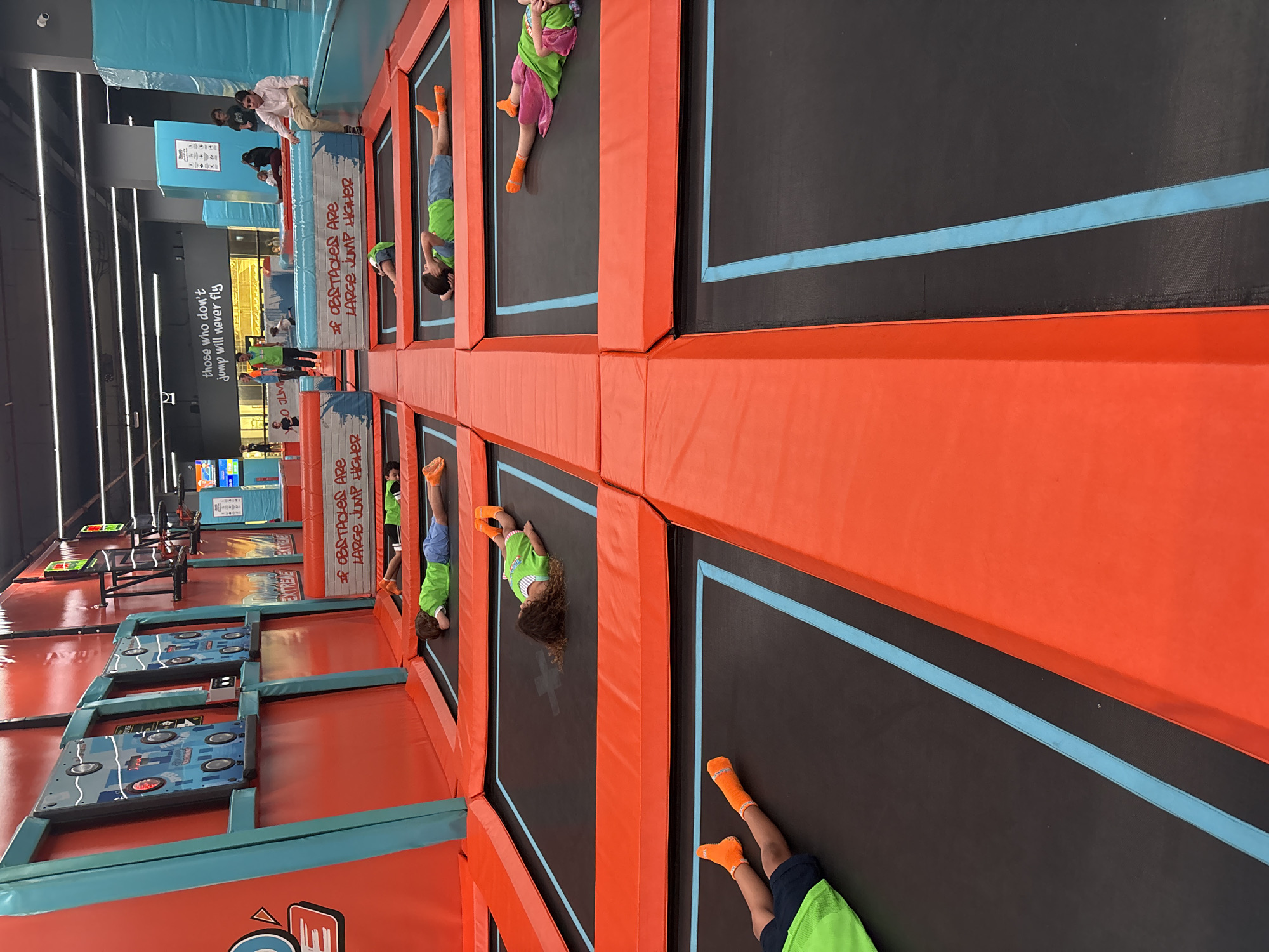 This image captures a wide view of an indoor trampoline park, likely located in Dubai, United Arab Emirates. The facility features numerous interconnected trampolines with black mats, each outlined by a light blue stripe and bordered by bright orange safety padding. Several children are visible across the trampolines in the foreground and mid-ground, mostly lying down in various resting postures. They are uniformly wearing bright orange grip socks and athletic attire, including green t-shirts and shorts in blue or pink.
In the background, padded walls are prominent, displaying a vibrant red and blue color scheme. On the left, a red wall features blue frames containing interactive game elements with illuminated targets. Text partially visible on these structures includes "RAMBO EXTREME". A white and blue padded barrier in the mid-ground prominently displays the slogan "IF OBSTACLES ARE LARGE JUMP HIGHER". Further back, a dark wall bears the white text "those who don't jump will never fly".
Several adults and other children are scattered in the background, either standing or sitting on various padded platforms and ledges. One adult male, dressed in a light shirt and trousers, is seated on a blue padded ledge on the right side of the image, observing the scene. The ceiling is dark with visible lighting fixtures, indicating a spacious, artificially lit environment. The overall impression is a vibrant, modern recreation center designed for active play.