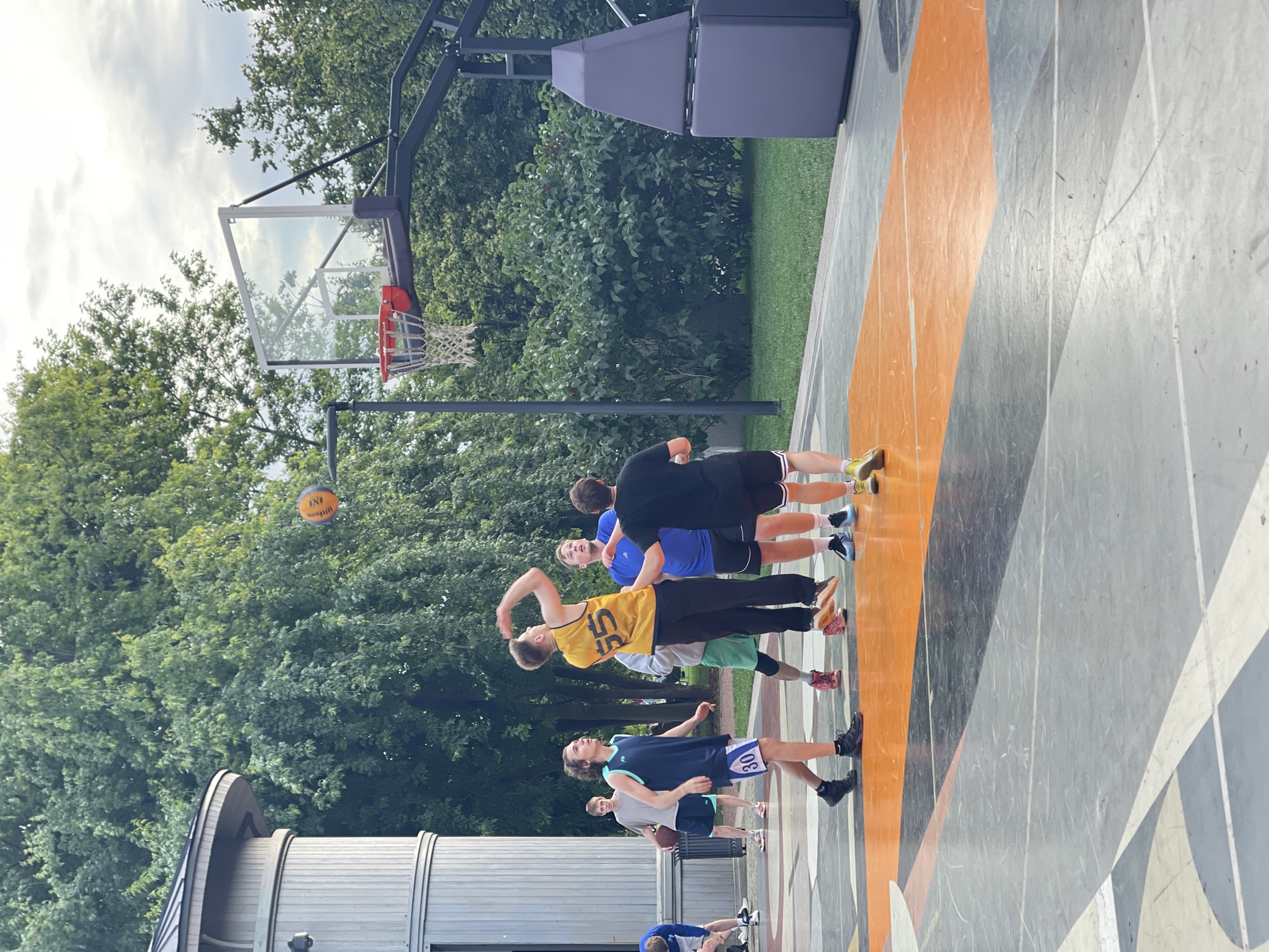 In an outdoor basketball court in Saint Petersburg, Russia, a group of young men are engaged in a game. The court features a black and orange surface with geometric markings. A regulation basketball hoop with a glass backboard stands prominently. Tall, leafy trees form a lush backdrop against a cloudy sky. One player in a yellow jersey with the number "55" is captured mid-air, attempting a shot. Other players, dressed in various athletic wear including a blue tank top with "30" on the shorts and a black t-shirt, are positioned around the court, some observing the play and others defending. A cylindrical building with a conical roof is visible to the left, partially obscured by trees. The overall scene depicts a casual, athletic activity in an urban park setting.