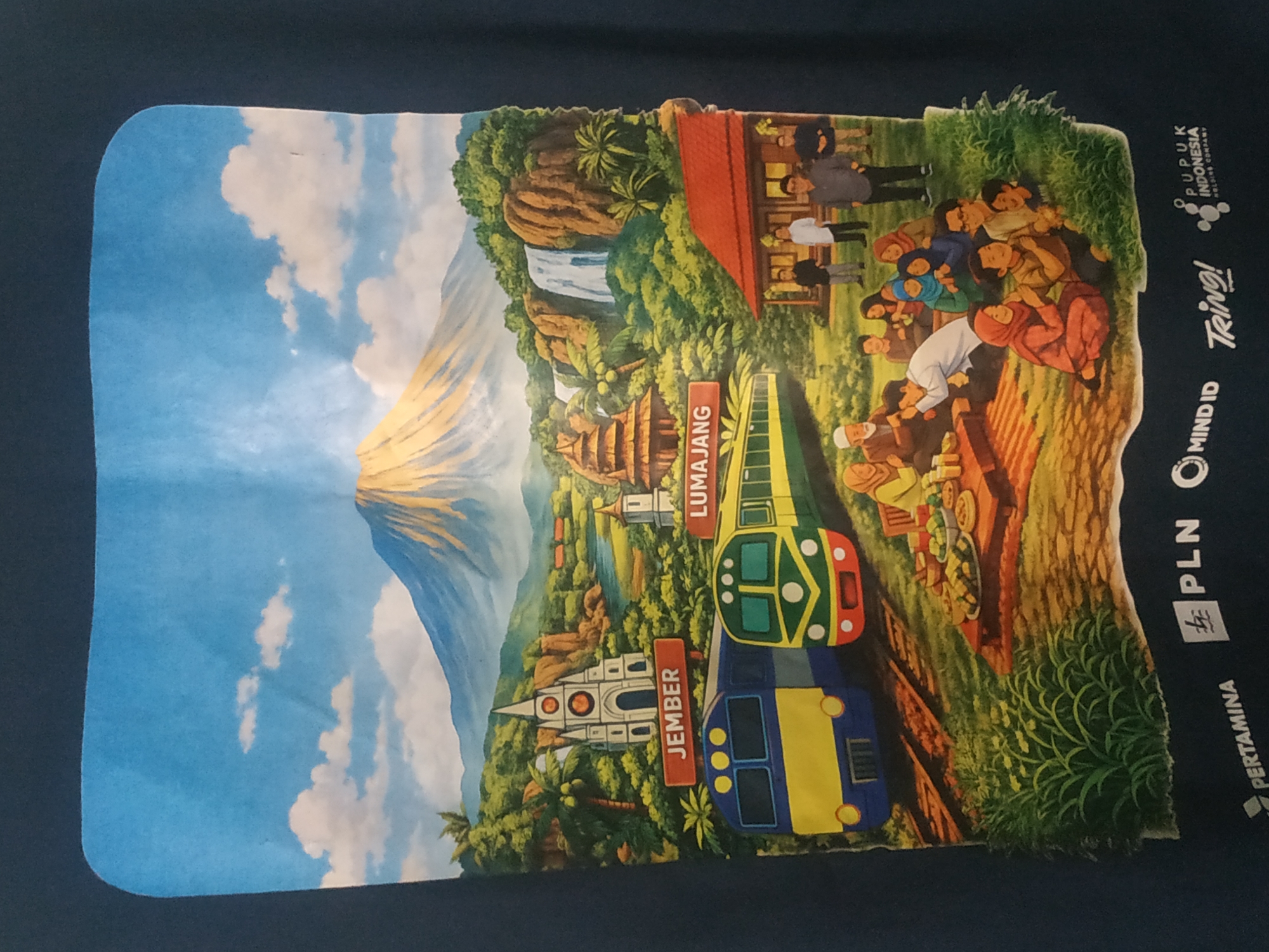 The image depicts a vibrant, stylized illustration of a landscape with a mountain, waterfalls, and lush vegetation, possibly representing the city of Arjasa, Indonesia. In the foreground, two trains are stopped on tracks. To the left, a blue and yellow train is parked next to a white building with a red-trimmed steeple. To the right, a green and yellow train is situated near a traditional Indonesian house with a red roof.
A group of people are gathered on mats laid out on the ground, enjoying a meal from a low table laden with food. Another group of people, dressed in formal attire, stand near the house. Several children are playing or interacting with adults in the scene. Signs indicate the locations of "JEMBER" and "LUMAJANG."
At the bottom of the illustration, several logos are visible: "PERTAMINA," a stylized lightning bolt, "PLN," "MIND ID," "Tring!", and "PUPUK INDONESIA HOLDING COMPANY." The overall scene evokes a sense of community, transportation, and local attractions within Arjasa.