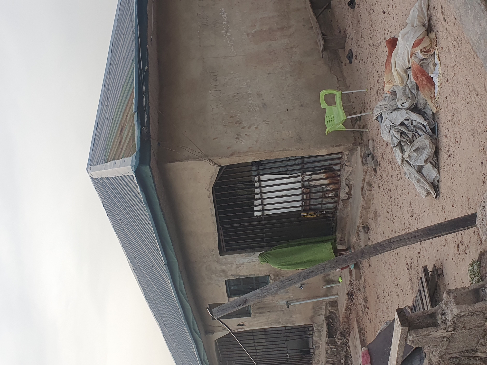 An outdoor shot depicts the side of a weathered, single-story building in Lokoja, Nigeria, under an overcast sky. The building features unpainted, rough concrete walls and a corrugated metal roof, possibly covered with a striped blue and white tarp or faded sheeting. Multiple windows are visible, all secured with dark metal bars. Behind the bars of an upper window, a person is partially visible, clad in a green garment, with their face obscured.
To the right of the building, a lime green plastic chair stands near a pile of assorted fabrics and clothing on the sandy, light brown ground. A long, dark wooden beam or plank extends diagonally across the mid-ground, passing below one of the windows. The ground in the foreground is bare earth with some scattered debris and what appears to be stacked wooden planks or furniture pieces. Electrical wires are strung along the building's exterior.
Visible text handwritten or spray-painted on the wall includes phrases like "I ACT-CALL", "5m 25 210/237-08MAE", and "2m KK / NZ 90", along with a partially visible "WET-CA". The scene captures a moment of daily life, with diffused daylight indicating either an overcast day or early/late hours without direct sunlight.