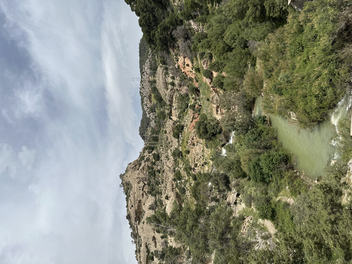 The image displays a rugged natural landscape in Ardales, Spain, centered on a deep valley or gorge. The sky, partially covered with scattered white clouds, fills the upper portion of the scene, with several power lines visible stretching across it and connecting to distant pylons on the mountain ridges.
Dominating the left and central areas are steep, rocky mountain slopes. These slopes are densely covered with various shades of green trees and shrubs, interspersed with exposed sections of light-brown and reddish-toned rock formations, indicating natural erosion.
Flowing through the lower right and extending into the mid-ground is a winding river with distinct greenish-tinted water. The river exhibits areas of faster flow, visible as white rapids and cascades, particularly towards the right side of the frame. Its banks are heavily forested with lush, green trees and thick undergrowth. Small, indistinct structures, possibly related to water management or infrastructure, can be faintly discerned within the vegetation near the river in the mid-ground.
No people are visible, emphasizing the natural and tranquil character of the location. The scene is illuminated by diffused daylight, suggesting an overcast or partly cloudy sky. There is no visible text in the image.