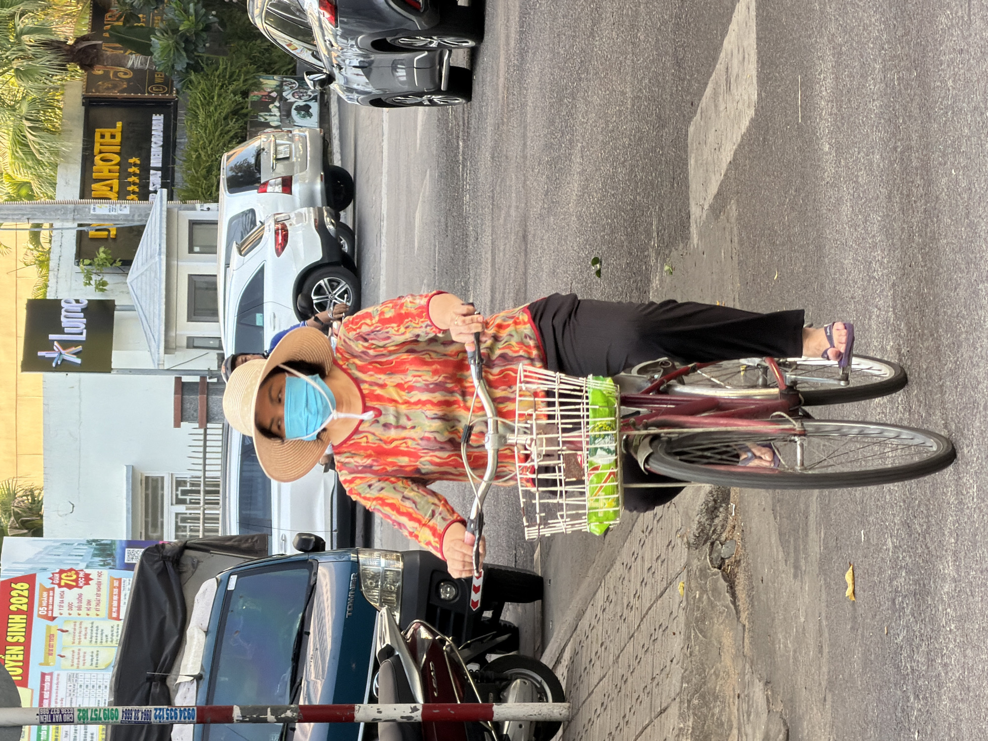 A woman wearing a wide-brimmed straw hat and a light blue face mask rides a red bicycle with a white basket on a street in Nha Trang, Vietnam. The basket contains several green bags, possibly snacks. She is wearing a colorful, patterned long-sleeved shirt and black pants. Her feet are clad in purple flip-flops. The bicycle's front wheel is turned slightly to the left, and the woman is looking forward. In the background, various vehicles are visible, including a blue truck, white SUVs, and a dark-colored car. A sign for "LUME" and "DLIA HOTEL" are prominently displayed on buildings. The street is paved, and there is a sidewalk with interlocking brick pavers to the left of the road. The environment appears to be an urban street scene.