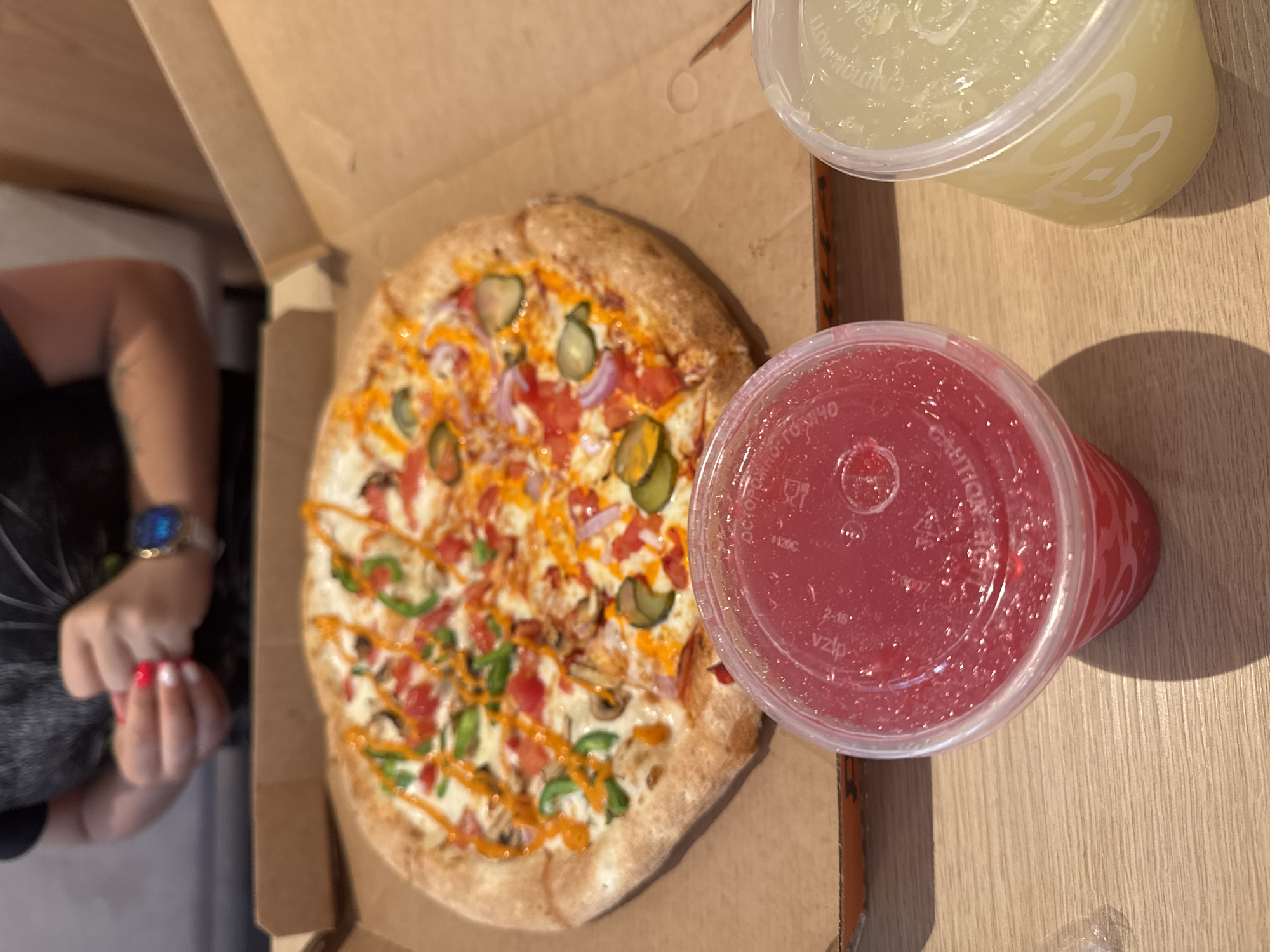 In Koltsovo, Russia, a pizza is presented in its cardboard box, situated on a wooden surface. The pizza is topped with tomatoes, green peppers, red onions, mushrooms, and a drizzle of orange sauce. In the foreground, two plastic cups are visible. The left cup contains a red beverage, and the right cup holds a yellow beverage. In the background, a person wearing a black t-shirt is partially visible, with their hands clasped and a watch on their left wrist. The environment appears to be an indoor setting, likely a restaurant.