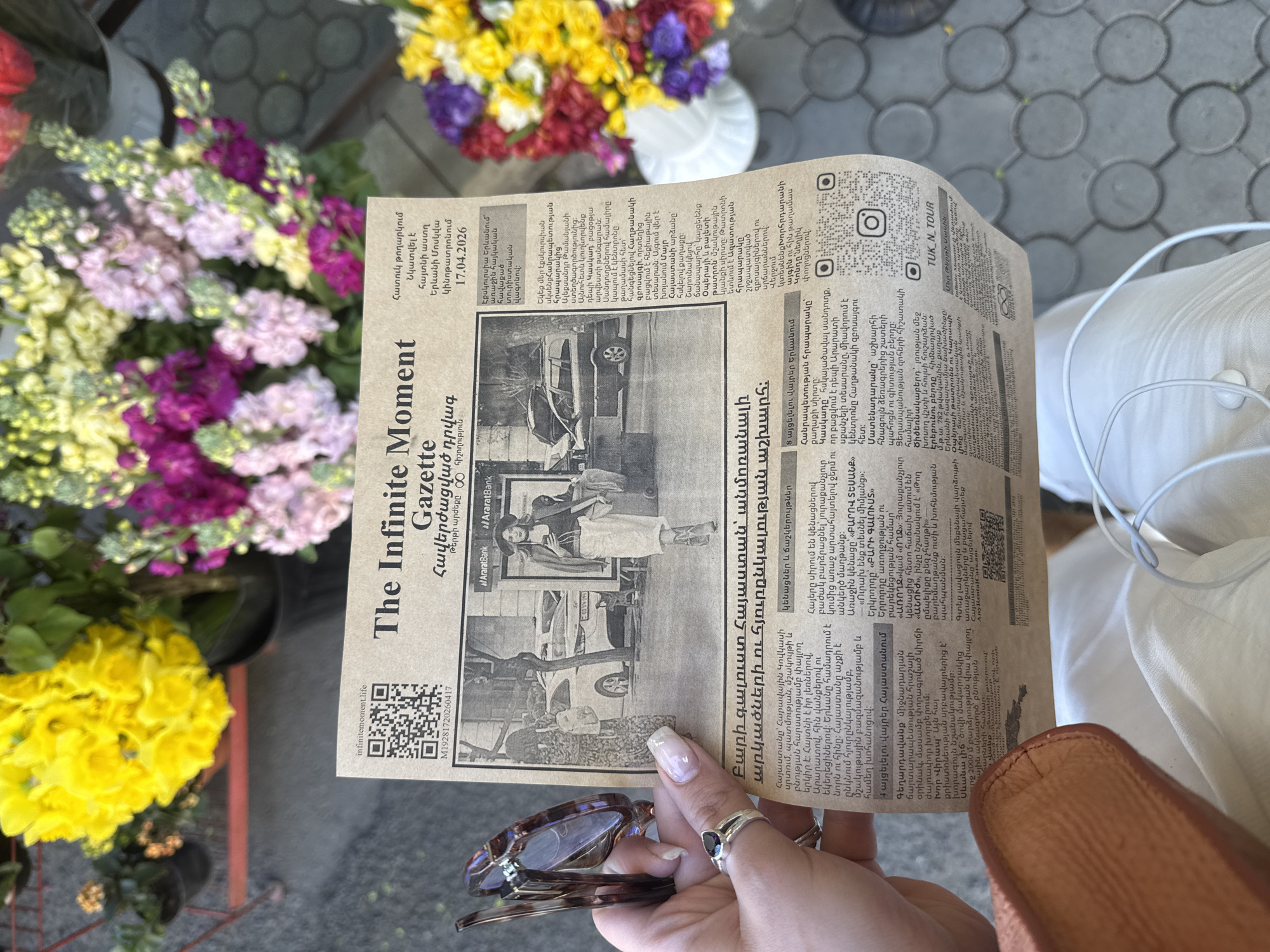 A close-up, high-angle outdoor shot captures a person's hand holding a folded newspaper amidst vibrant potted flowers and paving stones, likely in Yerevan, Armenia. The hand, adorned with polished nails and a ring featuring a dark stone, holds a brown tortoise-shell pair of sunglasses resting on the paper. The background features various colorful blossoms in shades of purple, pink, yellow, and white, suggesting a market or floral stall setting under bright daylight. A light-colored garment and white earbud cable are partially visible on the right.
The newspaper, titled "The Infinite Moment Gazette" with Armenian text translating to "Everlasting Episode" and "Newspaper price 100 memories," is a special issue. A prominent headline in Armenian welcomes readers to "Armenia – a world of unforgettable adventures and hospitality." Key articles mention a "Famous star spotted at Yerevan Moscow Cinema on 17.04.2026" and an "excursion in Yerevan with the first Armenian tourist wagon." It also details multiple Yerevan landmarks such as Republic Square, Cascade, Matenadaran, Tsitsernakaberd, Erebuni Fortress, Opera Theater, Parliament Garden, and the Kond district, alongside cultural insights into Armenian toasts and traditions.
Black and white photographs illustrate the content, one showing a person with a car, and another a couple. The newspaper features QR codes for infinitemoment.life, M19281720260417, @AraratBank, AnyRentalCars.am, and TUK_N_TOUR, along with Instagram logos. The publication functions as a promotional or cultural piece focused on Armenian tourism and local events, set in a sunny, pleasant outdoor environment.