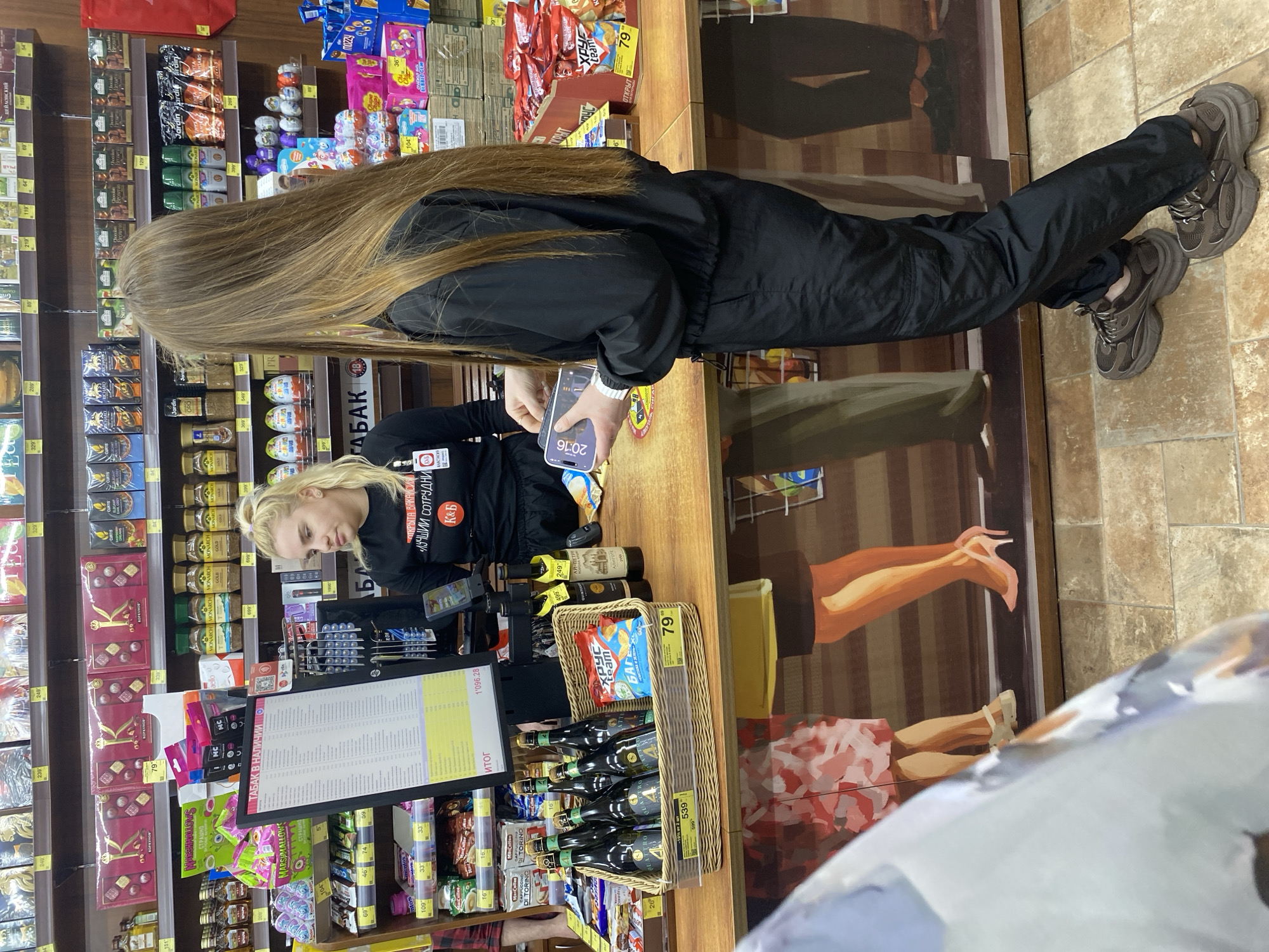The image shows a customer at a checkout counter in a store, potentially a К&Б (Krasnoe & Beloe) store, given the employee's shirt and the products visible. The customer, with long brown hair, is wearing a black tracksuit and large gray sneakers. She is using a smartphone with the time 20:16 displayed on the screen. A blonde cashier wearing a black shirt with Russian writing (Krasnoe & Beloe) is attending the checkout. Behind the counter, shelves are stocked with a variety of goods, including chocolates, coffee, and candies. A promotional display showing illustrated legs is attached to the counter. The counter also holds various products like wine bottles in a wicker basket and a bag of chips. A price list is visible on a screen in front of the cashier. The store seems to be in Yekaterinburg, Russia, given the Cyrillic text on the products and the employee's shirt.