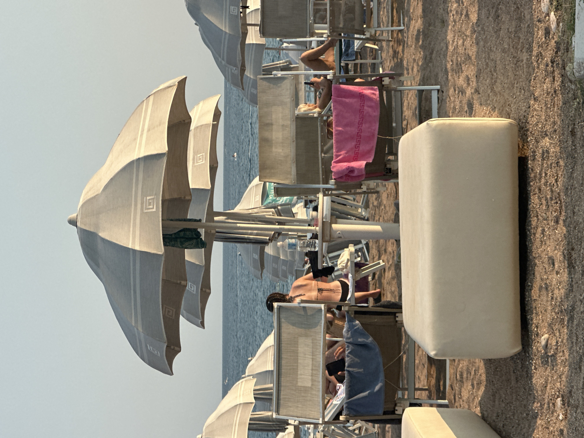The image depicts a beach scene in Boreti, Montenegro, with the Adriatic Sea in the background. In the foreground, two beige, rectangular ottomans are positioned on the sand. Behind them, several beach chairs and umbrellas are set up. In the middle ground, a woman in a black bikini is seated on a beach chair, facing away from the camera. Another woman is visible in a beach chair in the mid-right of the image, looking at her phone. The sea is calm with a few small waves. The sky is clear and pale blue. The overall atmosphere is one of relaxation and leisure on a sunny day.