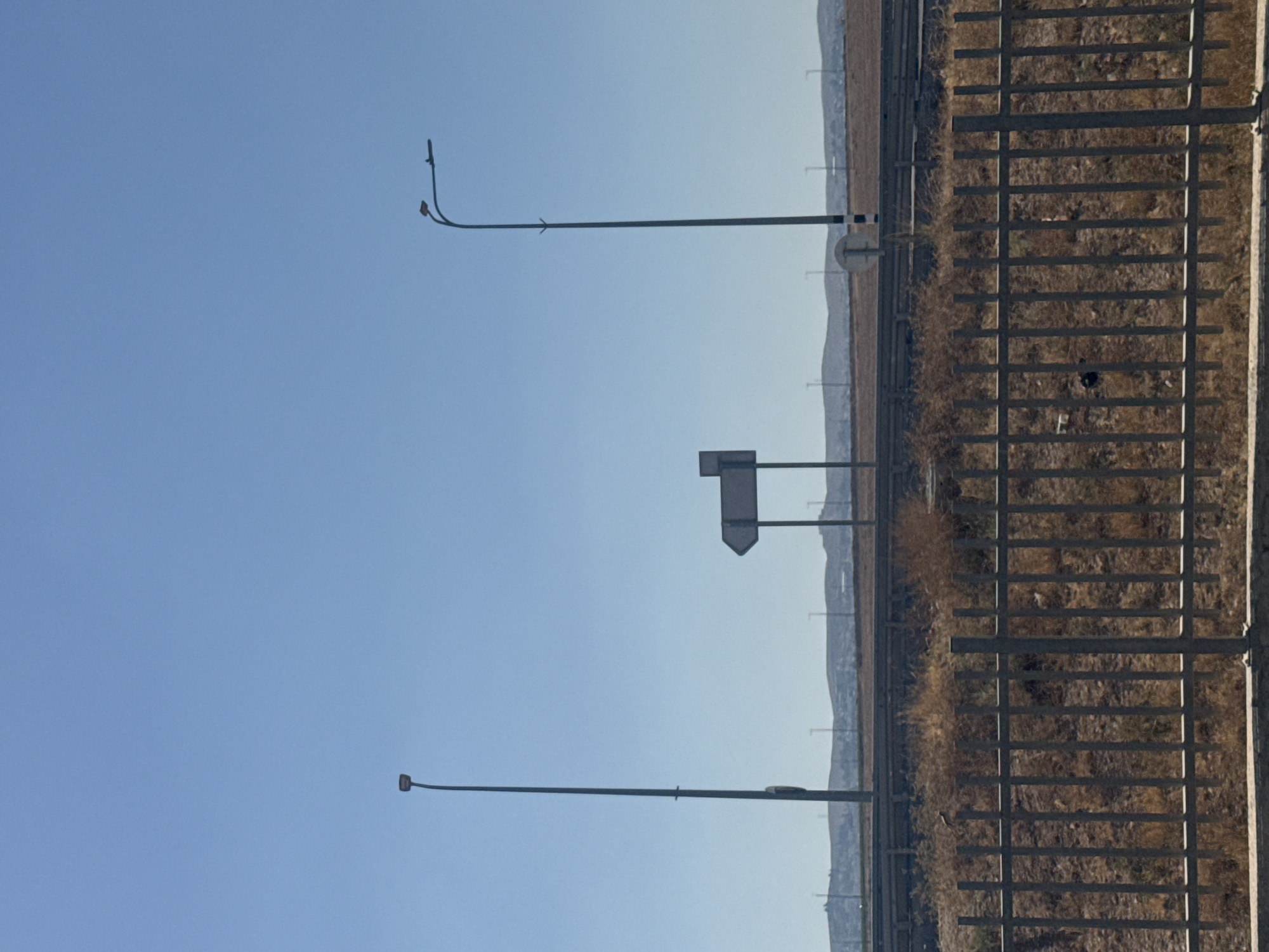 The image depicts a roadside scene with a clear blue sky and a dry, barren landscape. In the foreground, a metal fence runs horizontally across the bottom of the frame. Behind the fence, dry brush and vegetation are visible. Two tall streetlights stand on either side of the frame, with a directional signpost located between them. The signpost has a white rectangular sign with a black arrow pointing to the left. In the background, a series of hills and a distant horizon are visible, with scattered structures and more streetlights. The overall impression is one of a semi-arid environment, possibly along a highway or road in the Bnei Shimon Regional Council, Israel, characterized by its open spaces and sparse vegetation.