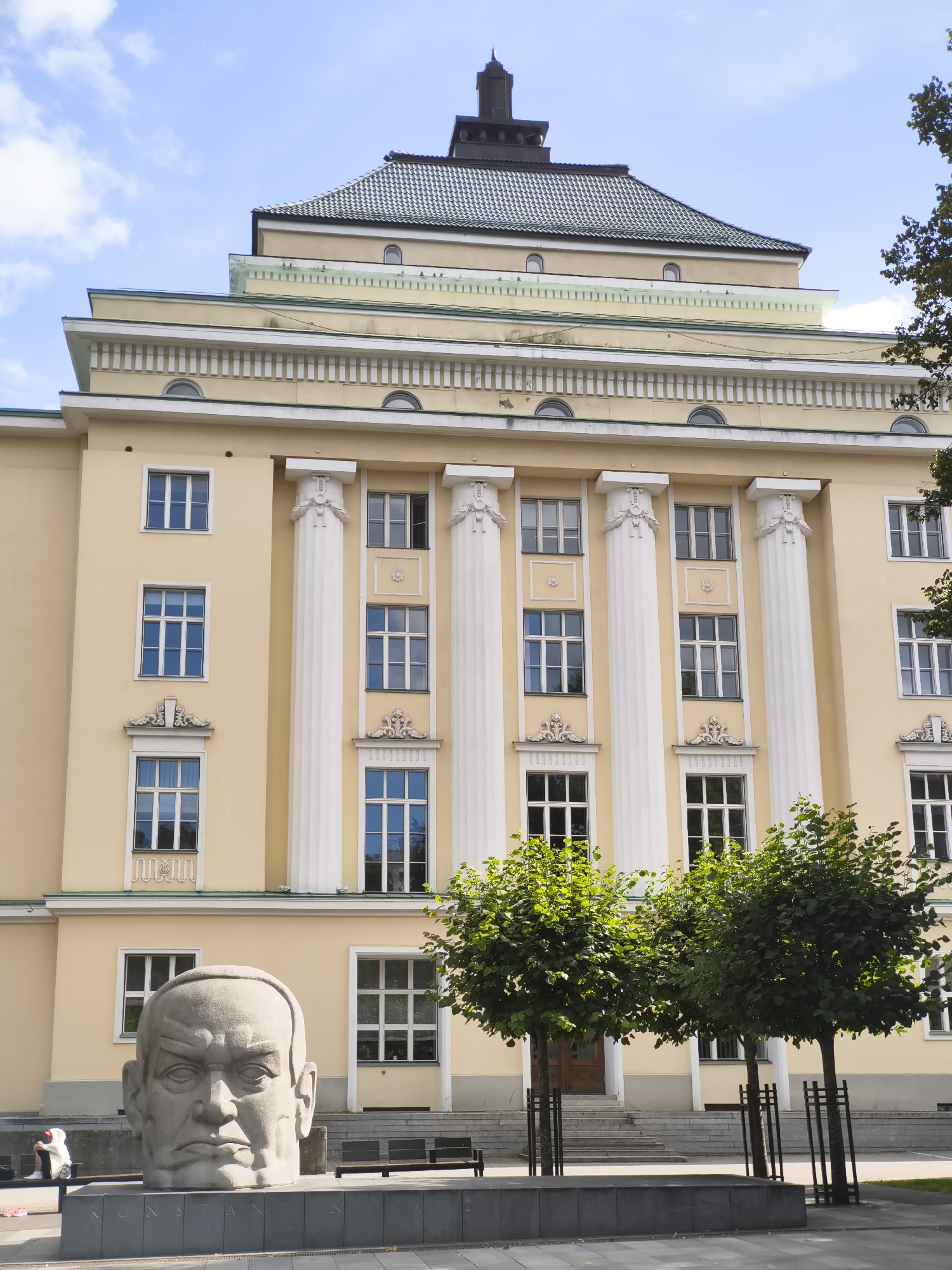 A large, light yellow building with a multi-tiered, dark green roof and a black spire is the primary subject. The building features classical architectural elements, including fluted white columns, ornate decorative elements above windows, and numerous symmetrically placed windows with white frames. In the foreground, a massive, gray stone sculpture of a man's head sits on a dark gray base with inscribed text. Two young trees with dense green foliage stand in front of the building, partially obscuring the lower part of its facade. The ground in front of the building is paved with gray stones. The scene is set under a bright blue sky with scattered white clouds. A person in white clothing is visible in the lower left corner, sitting on a bench near another person on a sidewalk. The building is located in Tallinn, Estonia, and the sculpture is of Konstantin Päts, the first head of state of Estonia.