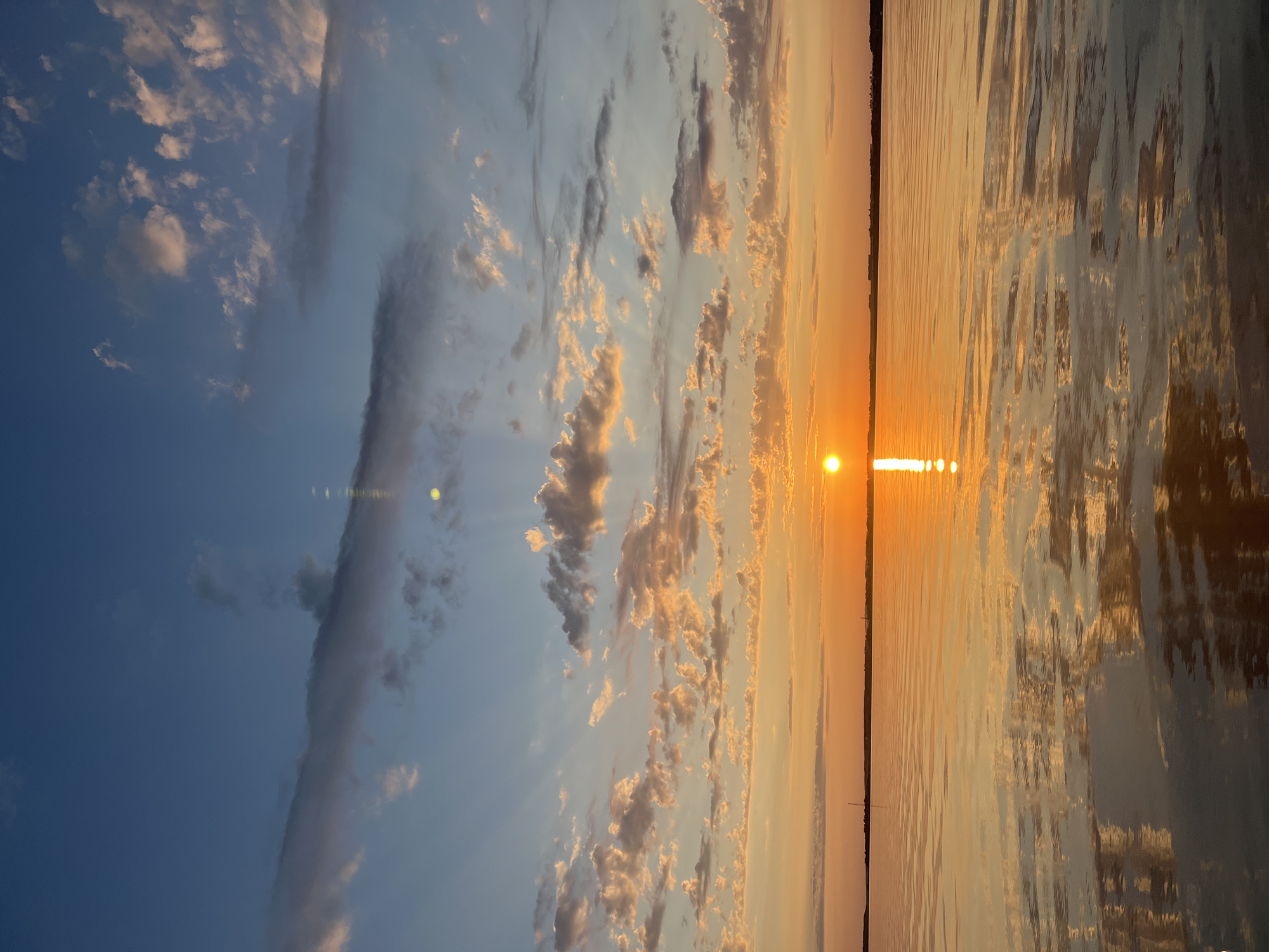 The image depicts a serene sunset over a body of water, likely a lake or river, near Novopichugovo, Russia. The sky is a gradient of deep blue at the top, transitioning to vibrant oranges and yellows near the horizon. Wispy clouds are scattered across the sky, catching the warm light of the setting sun. The sun itself is a bright, glowing orb positioned just above the horizon, casting a radiant reflection on the water's surface. The water is calm, with gentle ripples creating a shimmering effect. A thin strip of land is visible on the horizon, with subtle silhouettes of what appear to be buildings or structures. There are no people or animals visible in the scene. The overall atmosphere is peaceful and tranquil, characteristic of a sunset in a natural setting.