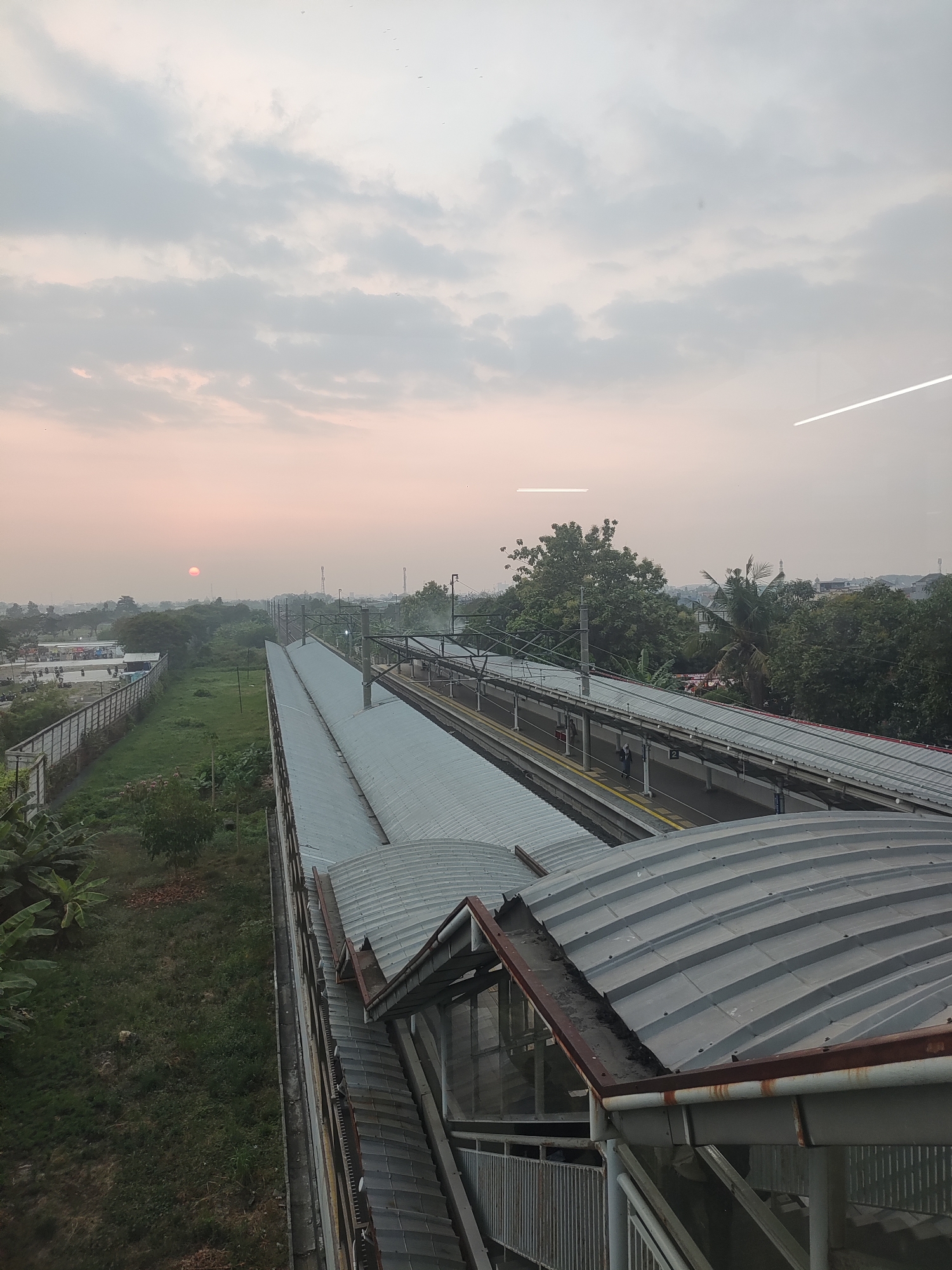 An elevated view captures a train station platform at Telagamurni, Indonesia, under a hazy, overcast sky with the sun appearing as a faint, reddish-orange disc on the horizon. The station features multiple parallel platforms covered by corrugated metal roofs supported by metal beams. A track runs between the platforms, with overhead electrical wires visible. Several people are seen standing on the platforms. To the left of the tracks, a grassy area borders a fence, beyond which are buildings and parked vehicles. Dense foliage and trees, including palm trees, surround the station and extend into the distance. The overall atmosphere suggests early morning or late afternoon lighting.
