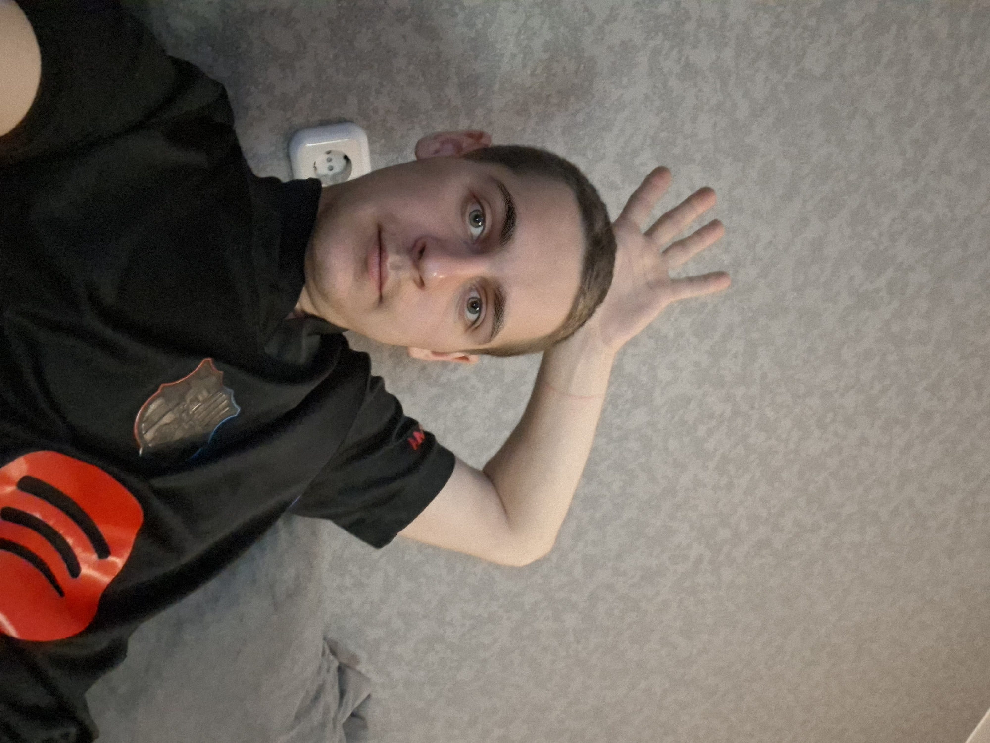 A person is lying down with a hand near their head, possibly on a bed or similar surface. The background is a patterned wall, with an electrical outlet visible near the person's head. The person is wearing a black shirt with a red logo and a badge on the chest. The location is Unknown, Unknown.
