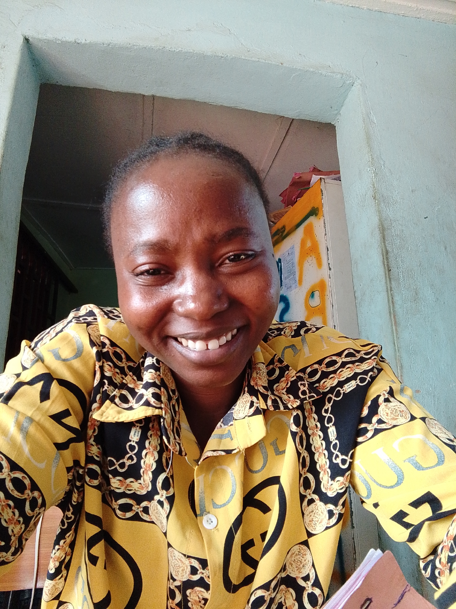A close-up, slightly high-angle shot captures a person smiling broadly into the camera, filling most of the frame. The subject is a young adult with dark skin and short, dark hair pulled back. A genuine, joyful smile reveals bright teeth, and the eyes are crinkled at the corners, conveying happiness.
The person is wearing a long-sleeved shirt with a distinctive pattern. The dominant colors are vibrant yellow and black, adorned with intricate gold chains, medallions, and stylized letters, including repeated 'U' and 'C' shapes. A white button is visible on the front of the shirt. In the bottom right corner, a brown object, possibly a book or folder, is partially visible, held by the person.
The background is an indoor setting, appearing to be a doorway or corner of a room. On the left side, a light gray/white textured wall is visible. On the right, the wall is painted a light blue/green, also textured. Behind the person's right shoulder, a colorful object, possibly a cabinet or storage unit, is visible. It features yellow and orange sections with graffiti-like markings, including a clear uppercase "A" in black and blue spray paint. The lighting is bright and natural, suggesting daytime. The overall emotion conveyed by the subject is one of cheerfulness and warmth.