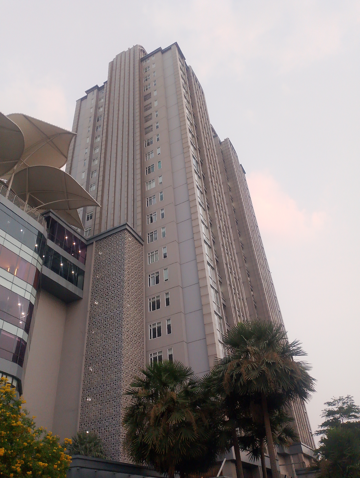The image shows a view of the high-rise apartments of Sudirman Park in Central Jakarta, Indonesia. The apartments are tall, grey buildings with many windows. In the foreground, there are palm trees and some yellow flowers. The sky is overcast.