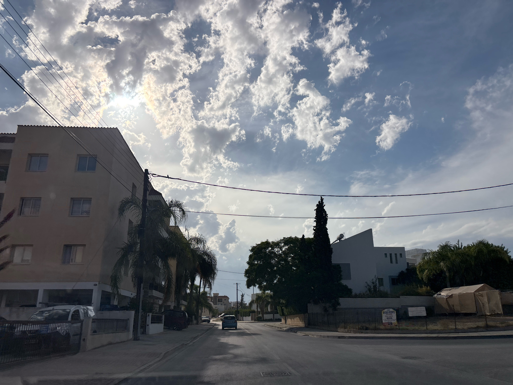 The image depicts a street scene in Mesa Geitonia, Cyprus, under a partly cloudy sky with the sun shining through the clouds. In the foreground, a paved road is visible, with a grey vehicle driving away from the camera in the distance. On the left side of the street, a light-colored, multi-story building stands with several windows. Palm trees are interspersed along the street. On the right side of the street, there are modern-looking white buildings, some with angular designs, and a large, dark green tree. A large tent or shelter covered in tan fabric is also visible on the right. Power lines crisscross the sky above the street. The overall impression is a residential or mixed-use urban environment.