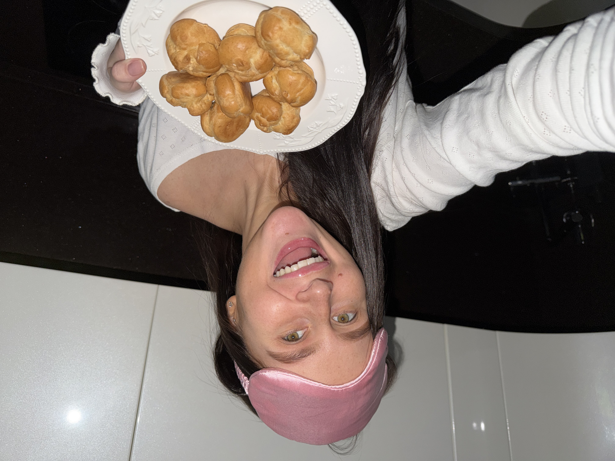 A woman is holding a plate of cream puffs in an upside-down selfie. She has long, dark hair and is wearing a pink eye mask on her forehead and a white, long-sleeved shirt. The background consists of dark countertops and white tiled walls. There is no verifiable information to definitively establish the location as Khimki, Russia.