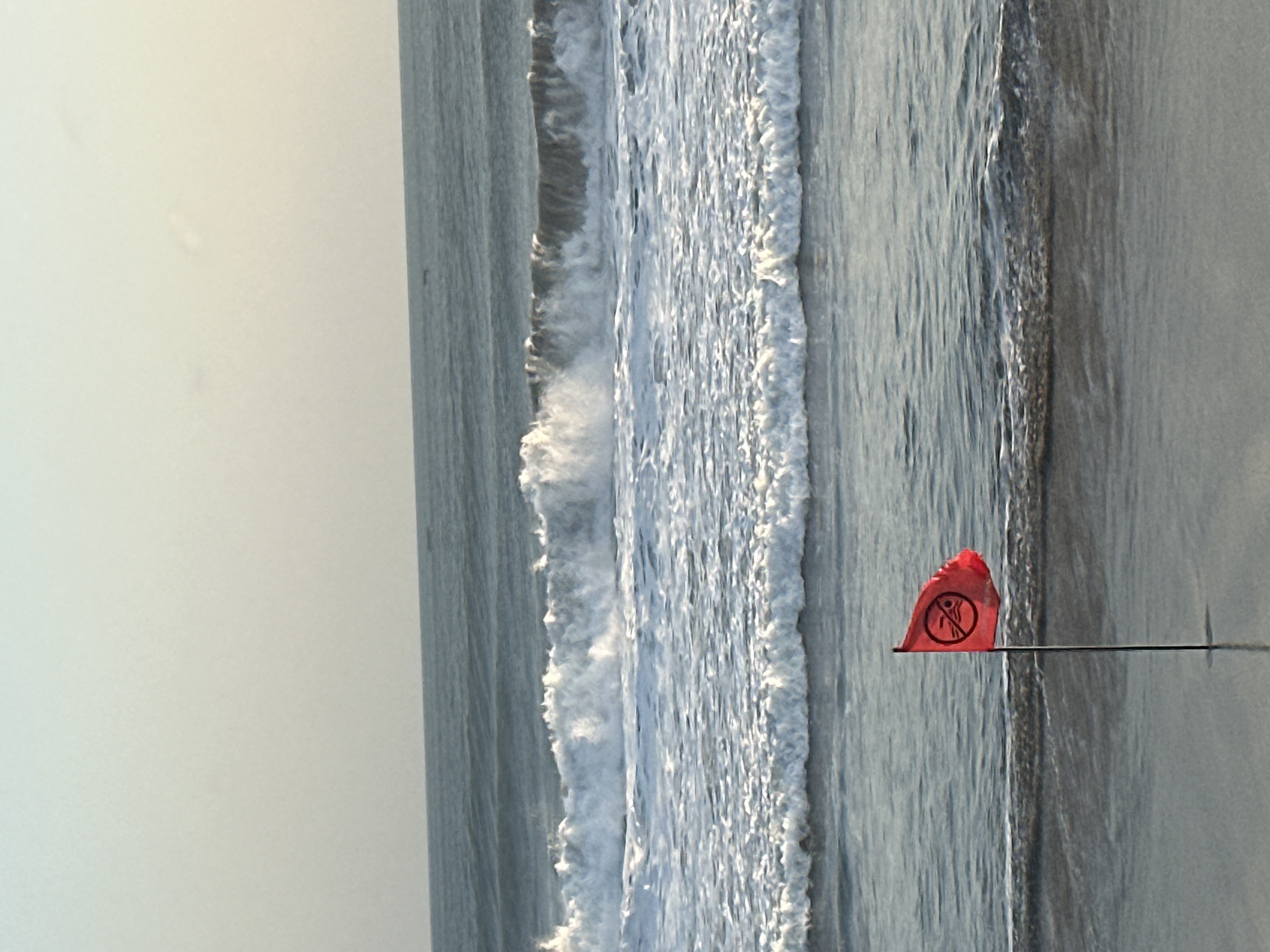 A red flag with a no-swimming symbol is planted in the wet sand at the edge of the ocean in North Kuta, Indonesia. The flag is positioned to the left of the frame, with its pole partially submerged in the water. Behind the flag, waves crash onto the shore, creating white foam. The ocean stretches out to the horizon, where the sky is a pale, hazy blue. The overall scene depicts a warning to beachgoers, suggesting that swimming conditions are not safe.