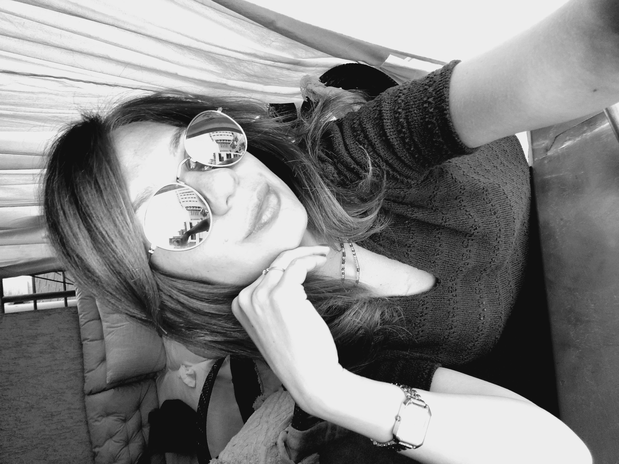 The black and white photograph shows a woman indoors, posing for a selfie. She is wearing large, round, reflective sunglasses, a V-neck top with a textured pattern, and a watch on her left wrist. Her right hand is resting on her chin, and she is looking into the camera. The background includes a wall with a textured finish, a bed with pillows, and a curtain. The reflection in her sunglasses shows a street scene with buildings. The image does not contain information about the location in Sofia, Bulgaria.
