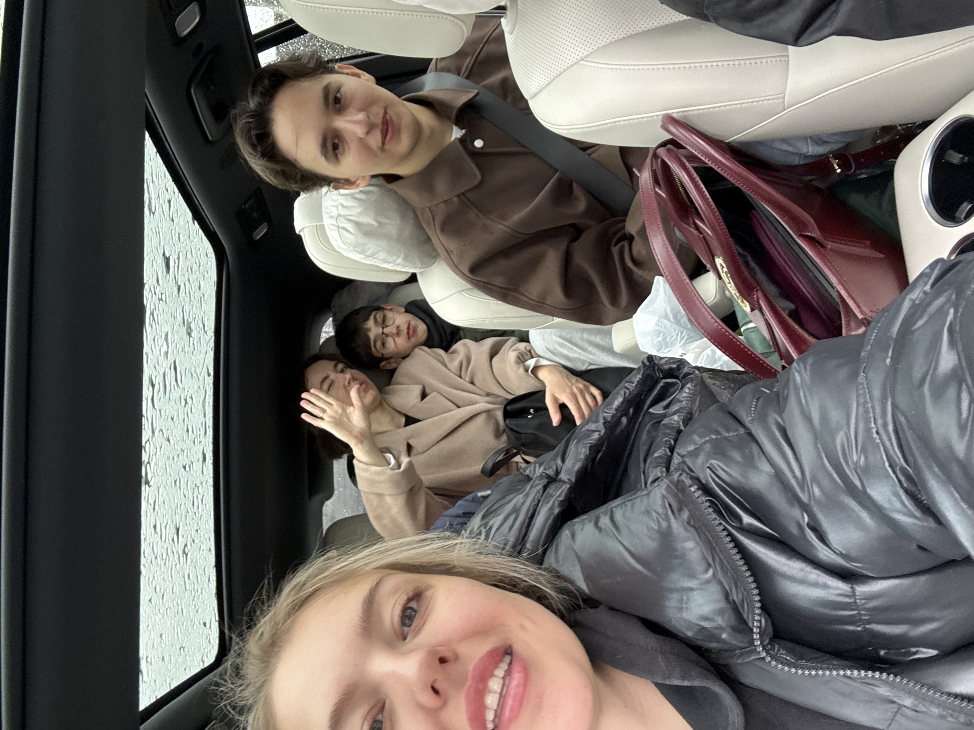 The image shows a selfie taken inside a car, likely in Солонцы, Russia, given the presence of a car and people who appear to be together. Four people are visible. The person taking the selfie is a blonde woman wearing a black puffy jacket. In the back seat, there are three other people: a man wearing a brown shirt and a seatbelt; two women laying down, one wearing a beige coat and waving, and another wearing glasses. A red purse and what appears to be bags and luggage are visible between the man and the backseat occupants. The window on the left has raindrops, suggesting it is raining outside. The car's interior has light-colored seats.