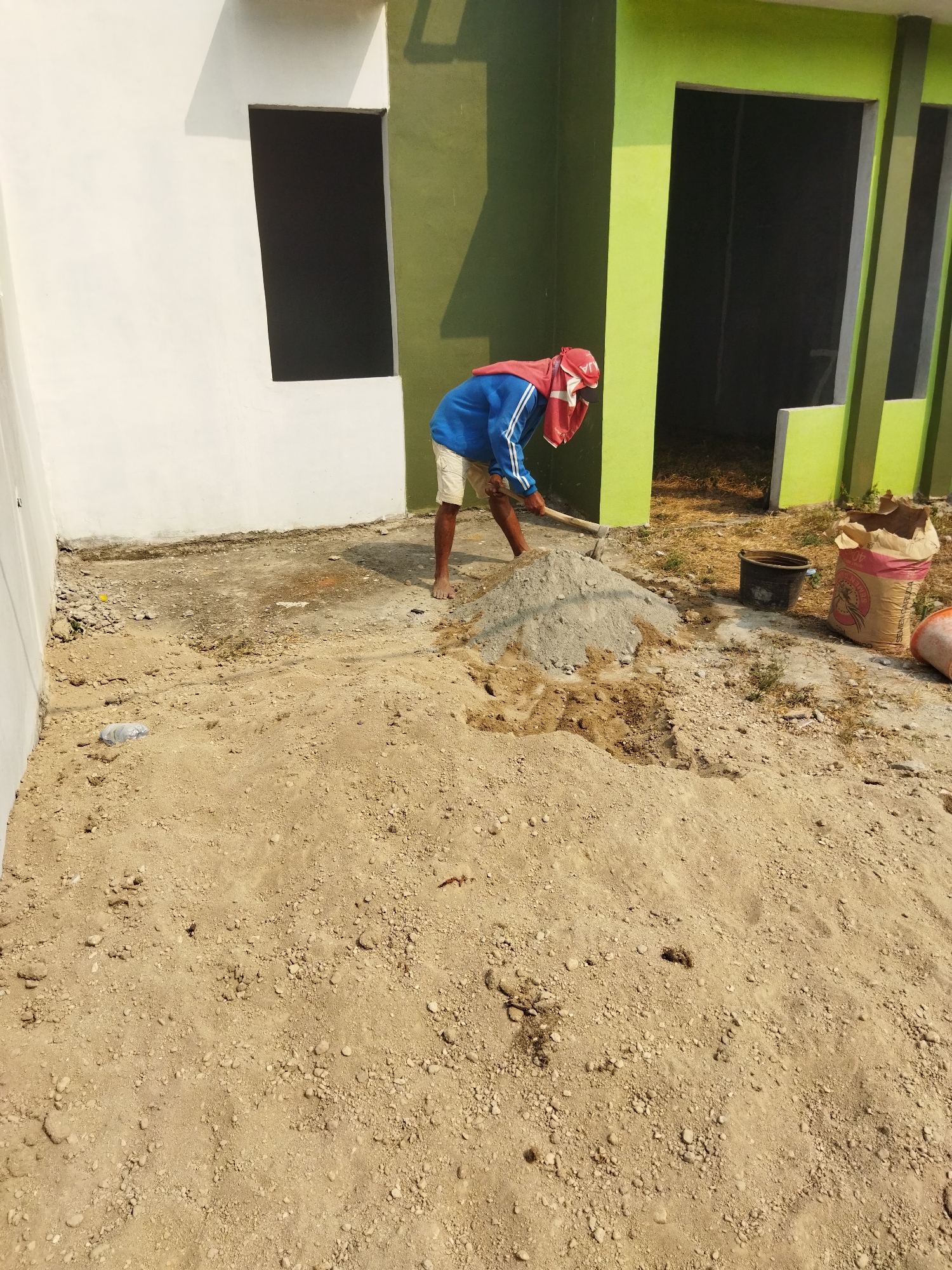 A construction worker in Suka Sari, Indonesia, is captured in a horizontal shot as they use a shovel to mix a pile of sand and cement. The worker, wearing a blue jacket with white stripes on the sleeves and light-colored shorts, has a red cloth draped over their head and shoulders for sun protection. They are positioned in the foreground, mid-action, with a large mound of mixed material in front of them. To the right of the mound, a black bucket and a half-full bag of cement are visible. The background features a building with white and lime green painted walls, and an open doorway. The ground is covered in dirt and some scattered debris. The scene appears to be an outdoor construction site under bright sunlight.