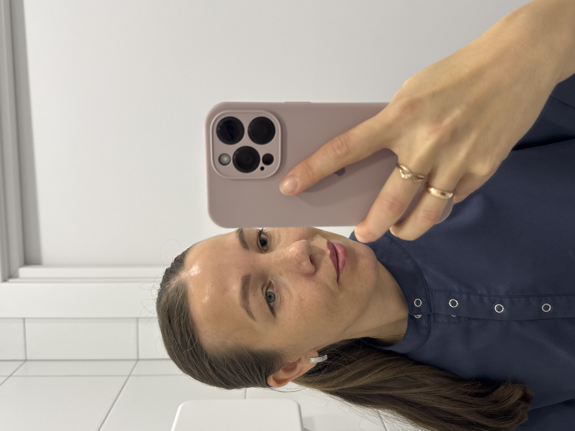 A woman, appearing to be in her 30s, takes a selfie in a brightly lit room. She has brown hair pulled back in a ponytail and is wearing a dark blue collared shirt with metallic button details. She has a light complexion, with some visible makeup on her face. She is wearing a silver hoop earring in her left ear and two gold rings on her right hand, which is holding a light pink phone. The phone has a prominent camera module with three lenses. The background appears to be a tiled wall on the left and a plain white wall on the right. The environment suggests an indoor setting, possibly a restroom or a clinical setting, given the sterile appearance of the walls. There are no visible indications of the city of Chaikovsky, Russia, in the image.
