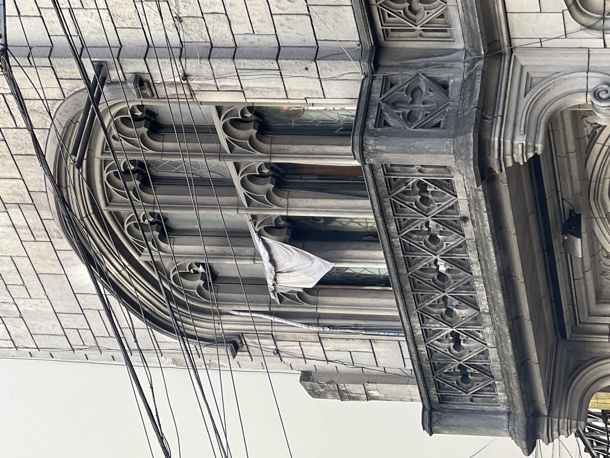 Low-angle shot of an ornate building facade with Gothic-style window arches and decorative stonework. The building appears to be located in Buenos Aires, Argentina. Numerous black electrical wires obscure parts of the facade. A single white cloth hangs from the building. The sky is a featureless white.