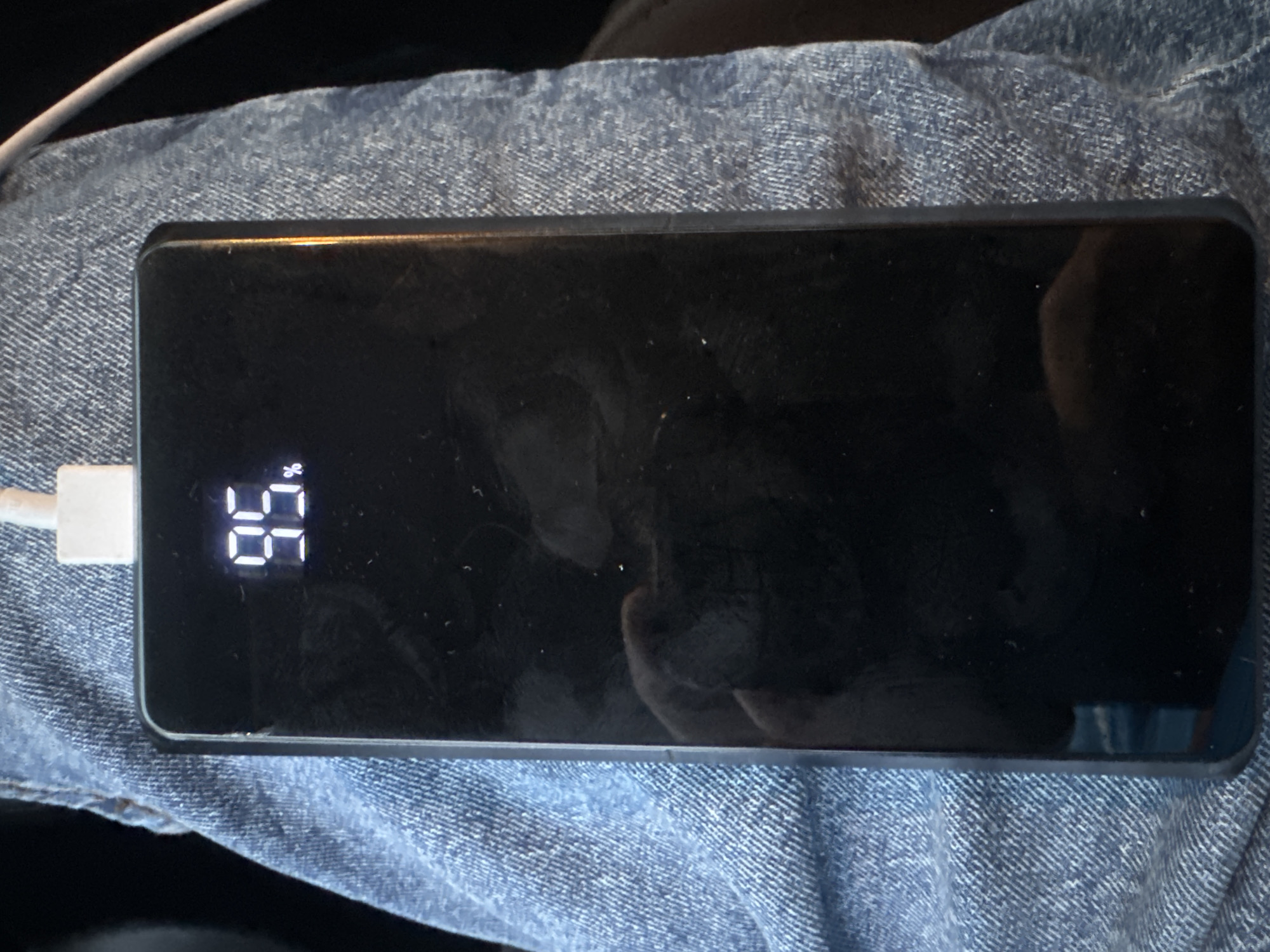 A black rectangular power bank is held in a person's lap, which is covered by blue denim jeans. The power bank has a digital display showing "95%" which indicates its charge level. A white charging cable is connected to the top of the power bank. The scene is dimly lit and there are reflections on the surface of the power bank, including a partial reflection of a hand. The background is dark and indistinct. There is no information available in the image to indicate the location in the city of Murcia, Spain.
