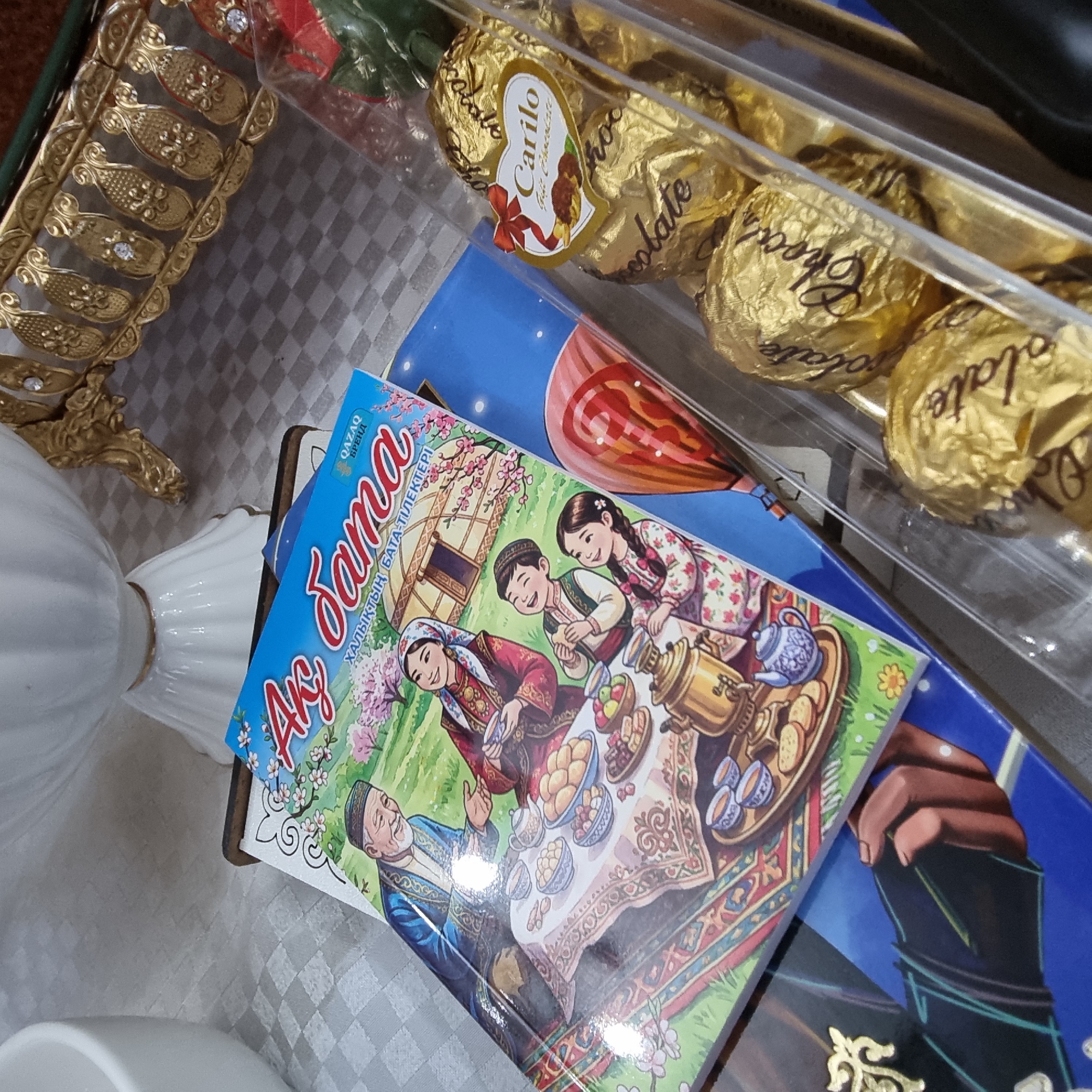 The image captures a tabletop scene, likely within an indoor setting in Aktobe, Kazakhstan. In the foreground, a clear plastic container holds several chocolates wrapped in golden foil. Each chocolate has the word "Chocolate" embossed in a cursive font. A red ribbon with a heart-shaped label that reads "Carilo" is visible on one of the chocolates.
Behind the chocolates, a book with a blue cover is partially visible, featuring an illustration of a family enjoying tea around a table laden with food. The illustration depicts traditional Kazakh attire and a yurt in the background, suggesting a cultural theme. The book's title, "Ақ Сама" and "ҚАЗАҚ БРЕНД", are visible in Kazakh script.
Further in the background, a decorative golden object with intricate patterns and small jeweled accents is visible, possibly part of a tray or display. A white porcelain object, perhaps a vase or bowl, stands behind the book and decorative item. The surface the objects are placed on appears to be a checkered tablecloth. The overall impression is of a domestic setting, possibly a gift presentation or a display of cultural items.