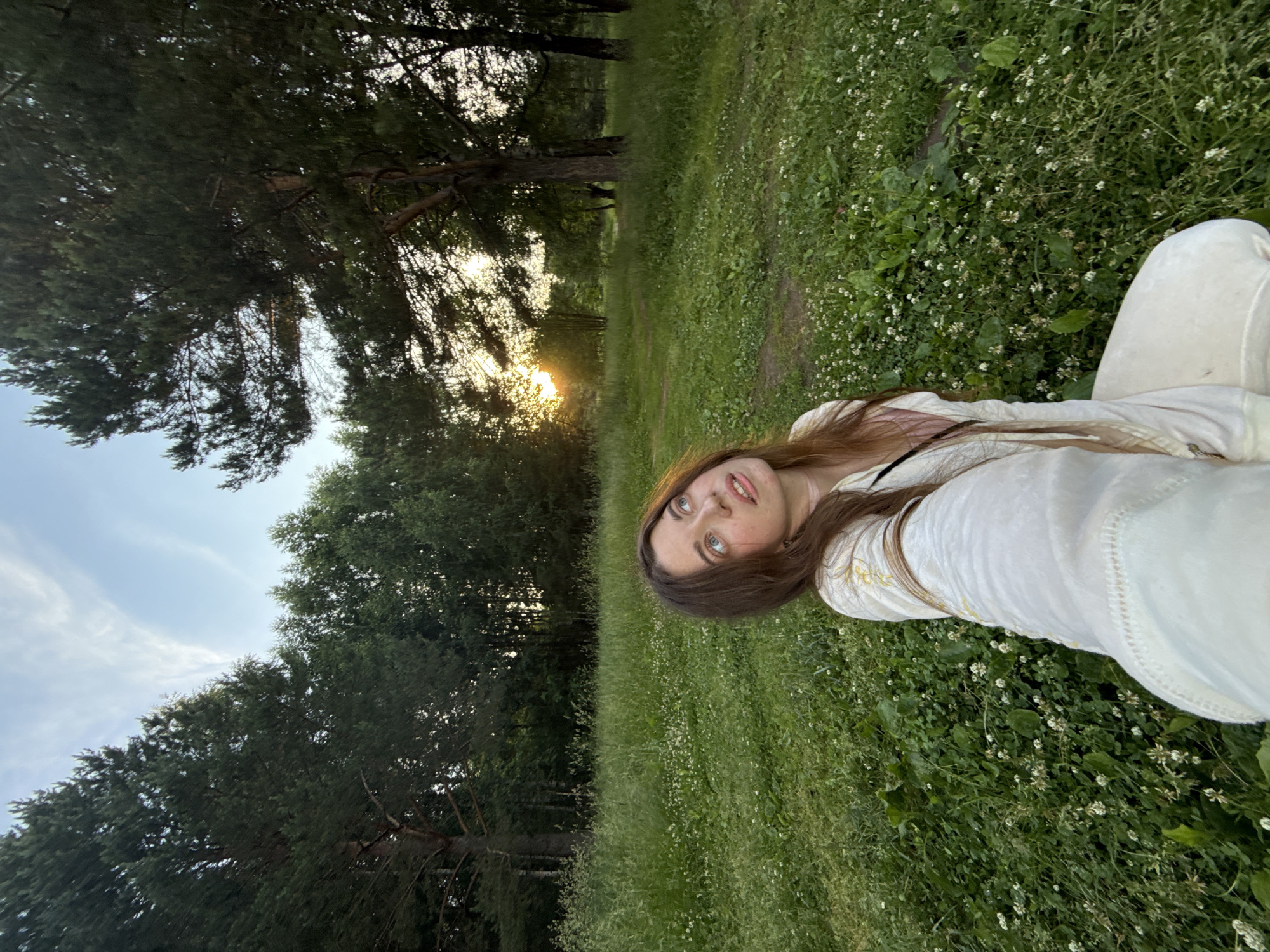 An outdoor, low-angle, eye-level selfie taken by a woman sitting in a field of grass with scattered white flowers. She has long, brown hair and is looking up and to her right with her mouth slightly open. She is wearing a white outfit with yellow embroidery on the sleeves and a pink top underneath. The background consists of a line of trees, some of which are tall pines, against a pale blue sky with wispy clouds. The sun is setting behind the trees on the left side of the image, casting a warm glow.