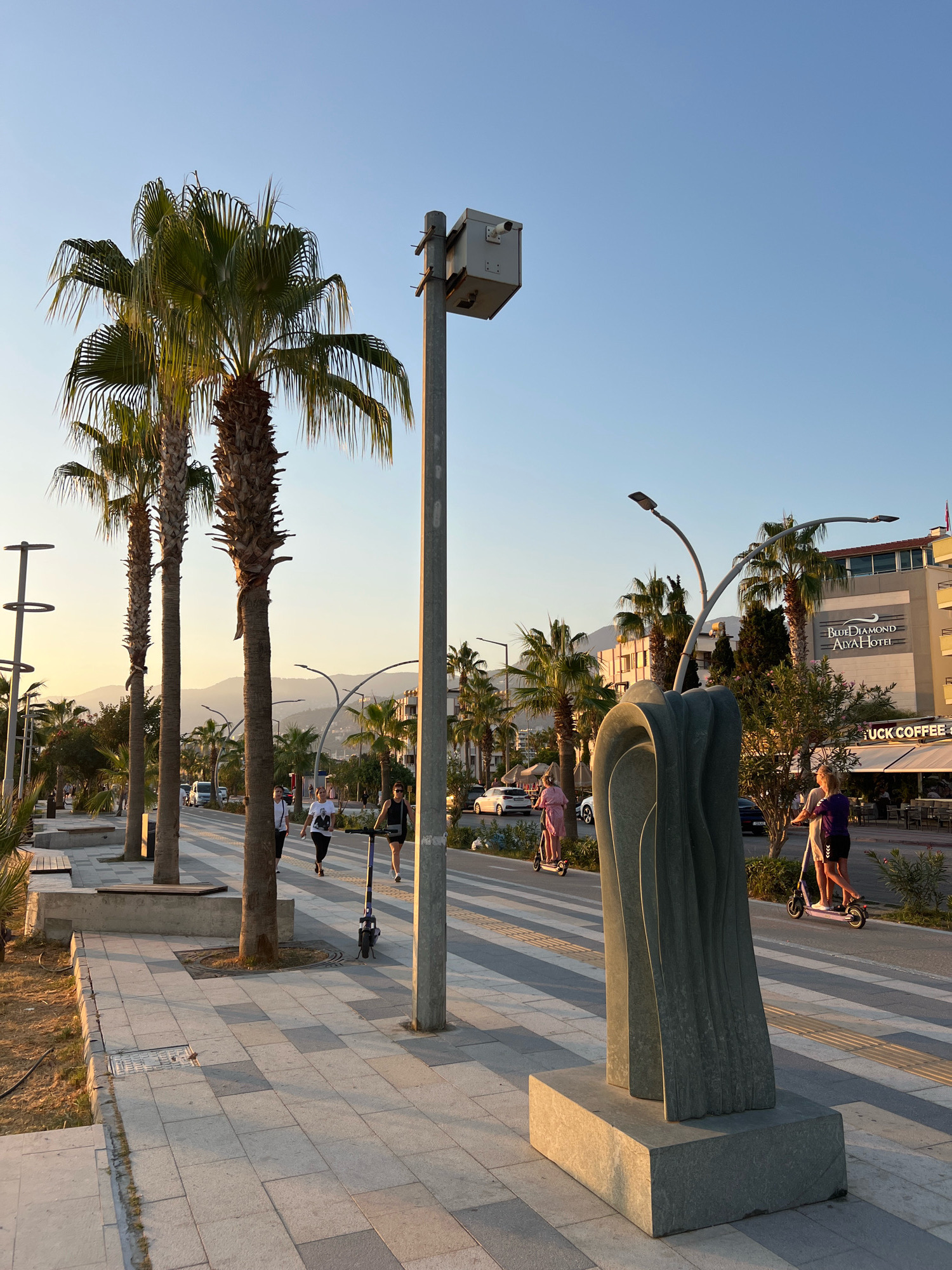 The image depicts a street scene in Alanya, Turkey, during what appears to be late afternoon or early evening, indicated by the warm, golden light casting long shadows. The foreground features a wide, paved pedestrian walkway with a striped pattern. A modern, abstract sculpture made of stone stands prominently on the right side of the walkway. Beyond the walkway is a street with light traffic, including cars and electric scooters.
Several palm trees line the left side of the walkway, providing shade. In the background, a multi-story building with "Blue Diamond Alva Hotel" signage is visible, along with other businesses like "UCK COFFEE." The sky is clear and blue, with distant mountains faintly visible on the horizon.
People are engaged in various activities. Two individuals are riding electric scooters on the street. Further down the walkway, a woman in a pink dress is also on an electric scooter. Two other individuals are walking on the walkway, one in a white t-shirt and the other in a dark top. A woman in a purple top is also seen on an electric scooter near the sculpture. A street lamp with a box-like fixture is positioned behind the sculpture. The overall atmosphere is that of a pleasant, active urban environment by the coast.