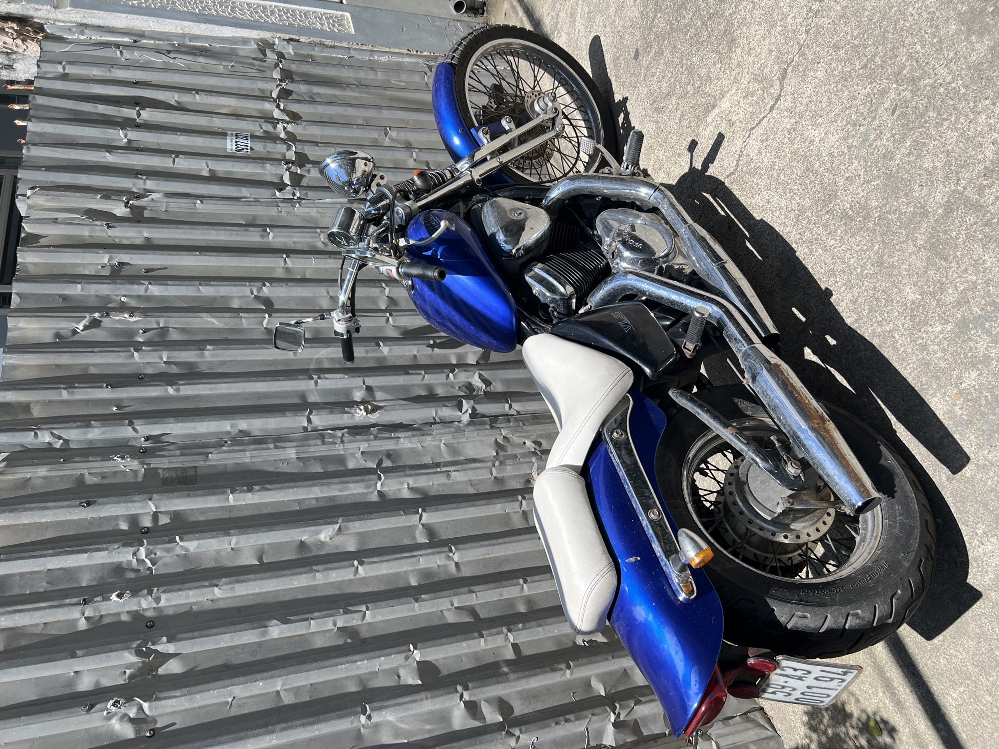 The scene is located in an urban area of Nha Trang, Vietnam, featuring a blue motorcycle parked on a concrete surface against a corrugated metal fence.
The primary subject is a metallic blue Honda Shadow VLX (model designation "VLS" visible) cruiser-style motorcycle, registered with license plate "59-A3 001.94", indicating Ho Chi Minh City registration. The motorcycle features a two-part white seat with purple stitching, extensive chrome components including handlebars, mirrors, headlight, engine covers bearing the "Honda" logo, and twin exhaust pipes. The front wheel has wire spokes, while the rear wheel features a solid disc hub with spokes. Both tires exhibit moderate tread wear.
In the background, a heavily corrugated, galvanized steel fence or gate stands vertically, exhibiting numerous dents and localized surface rust. The fence is secured by visible fasteners. A small white rectangular sticker with black text, including the partial phone number "0937 287", is affixed to the corrugated surface. To the right of the fence, a section of a light-colored building wall with a textured, decorative pattern is visible. A dark, cylindrical pipe runs vertically along the extreme right edge of the frame.
The motorcycle is stationary, parked upright on a light grey concrete pavement that displays minor surface cracks. Strong, direct sunlight illuminates the scene from the upper left, casting distinct, well-defined shadows of the motorcycle and its components onto the concrete. No people are present in the image.
